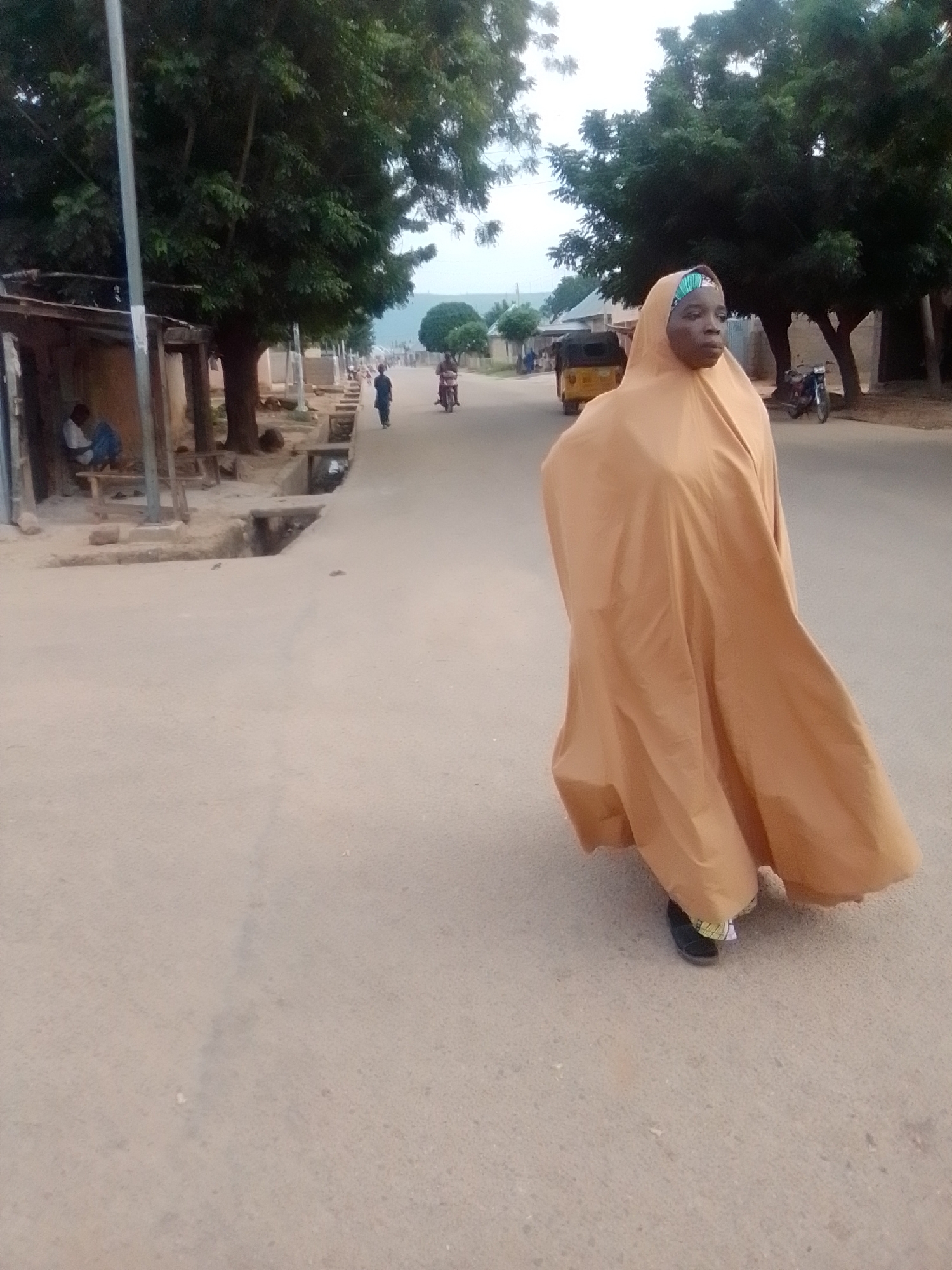 A woman walks on a paved road in Liji Hill, Nigeria, wearing a tan hijab and a matching flowing garment. She is partially obscured by her clothing, but her face is visible. In the background, a yellow auto-rickshaw is visible, along with a motorcycle and several trees lining the road. To the left of the road, a small structure with a thatched roof is present, and a person is seated inside. The sky is overcast, suggesting it may be early morning or late afternoon.