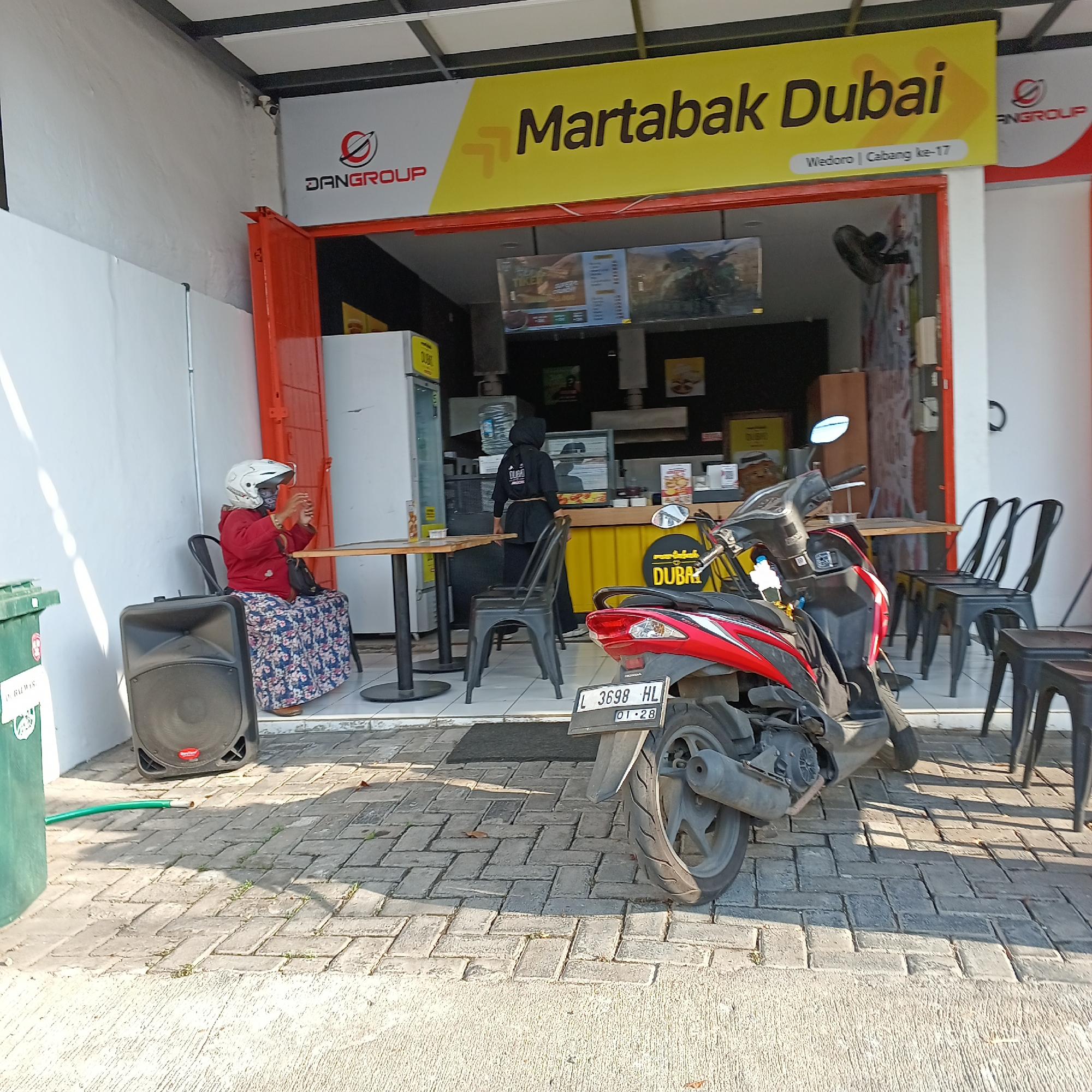 An outdoor street-level shot shows the exterior of a "Martabak Dubai" establishment in Wedoro, Indonesia. The facade features a large yellow sign with the words "Martabak Dubai" in bold, dark lettering. Above this, a smaller sign reads "DANGROUP" with a circular logo. A person wearing a white helmet, a red jacket, and a floral patterned skirt is seated at a small table to the left, looking at their phone. A large black speaker is positioned on the ground next to them. A red motorcycle is parked in front of the establishment to the right, with its license plate visible, reading "L 3698 HL 01-28". Several black chairs and tables are arranged outside the shop. A green trash can is visible on the far left. The ground is paved with brick pavers. The overall scene is well-lit by sunlight, casting shadows.