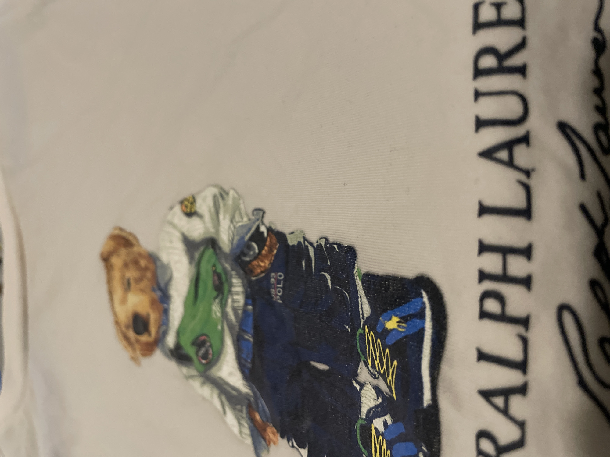 The image shows a portion of a white T-shirt with a Ralph Lauren Polo Bear design printed on it. The design features the Polo Bear wearing a racing suit. The text "RALPH LAUREN" is printed vertically alongside the image. There is no discernible information to suggest this image was taken in Barcelona, Spain.