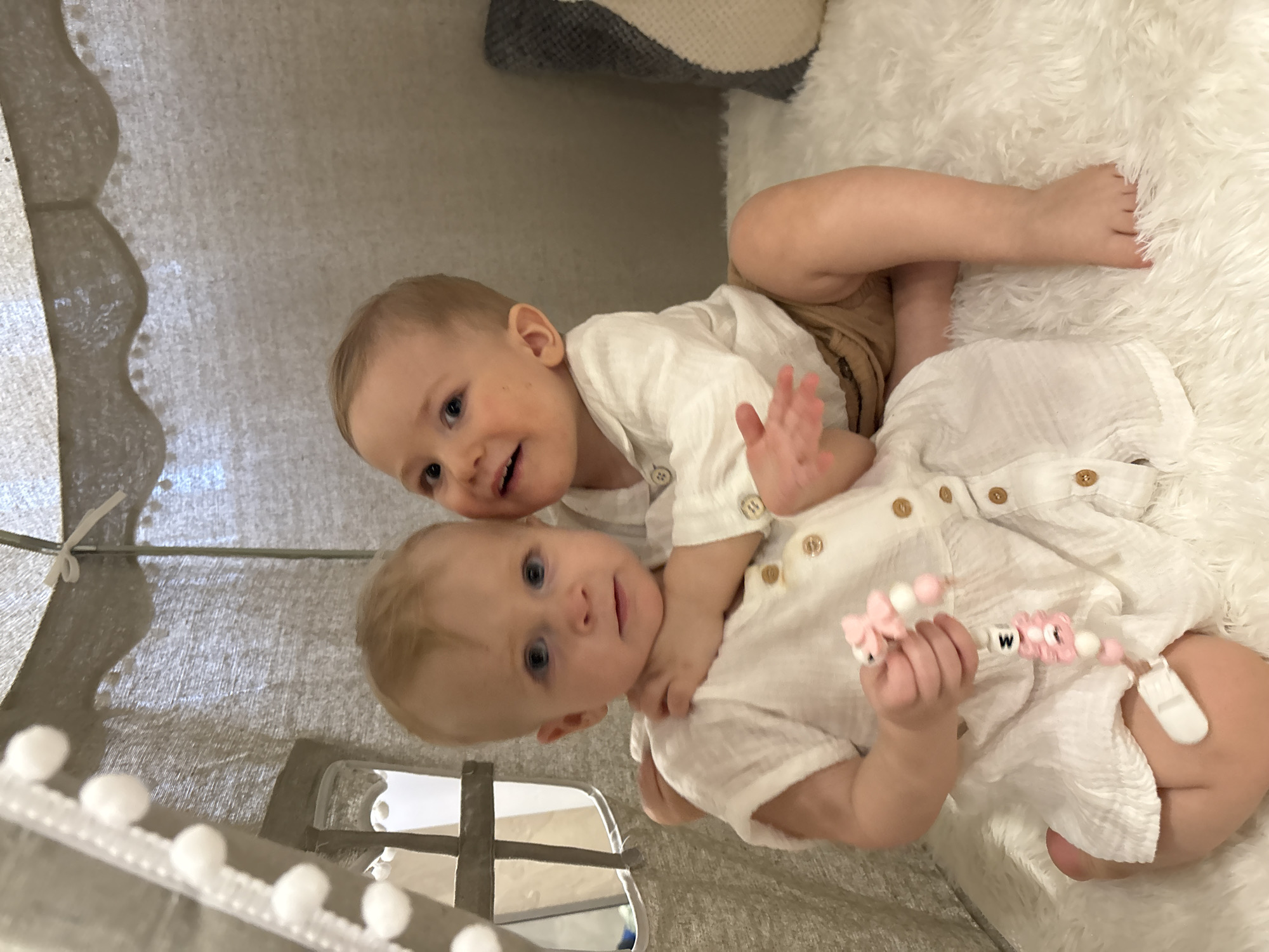 Two infants are playfully interacting within what appears to be a children's play tent. One infant is lying down wearing a white buttoned shirt, holding a pink pacifier clip with the letter "W". The other infant is sitting on top, also wearing a white buttoned shirt and tan shorts, with their right arm raised in a waving gesture. The background includes a fluffy white surface and part of the tent structure with pom-poms. It is not possible to confirm any location or city details.