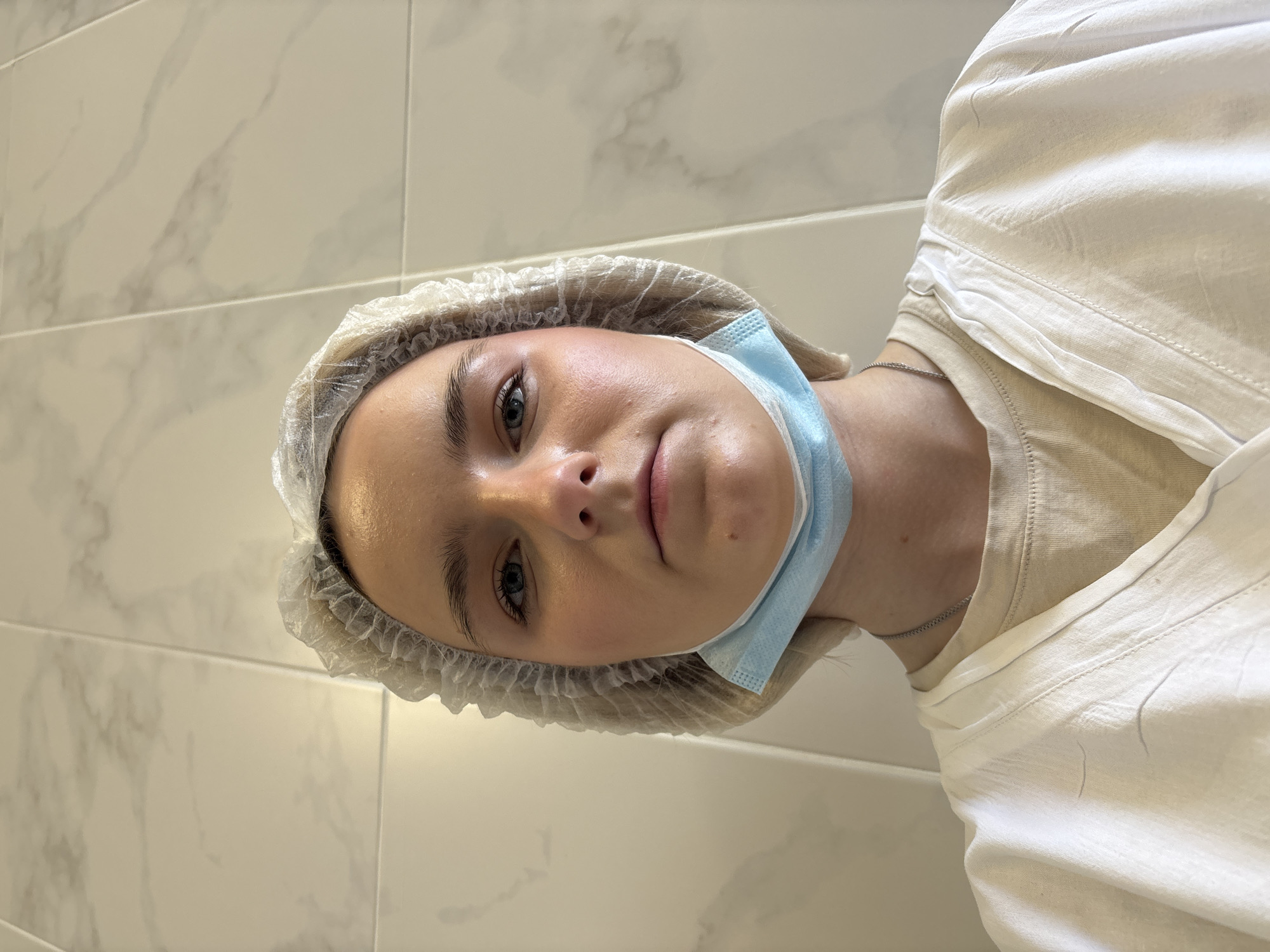 A close-up, upward-angled portrait features a young individual, likely female, with fair skin, light-colored hair, and light eyes, looking directly at the camera with a neutral expression. The setting appears to be indoors, possibly in Saint Petersburg, Russia, against a wall clad with light-colored, marble-patterned rectangular tiles.
The subject wears a disposable, translucent bouffant cap over their hair and a light blue surgical mask pulled down below their chin. They are dressed in a light-colored (possibly beige or off-white) t-shirt, over which a loose-fitting, lighter-toned garment, such as a smock or gown, is worn. A thin silver chain necklace is visible around their neck. The lighting highlights their skin with a slight sheen. The overall impression is calm, suggesting a moment either before or after a procedure in a clinical, aesthetic, or sterile environment. There is no visible text in the image.