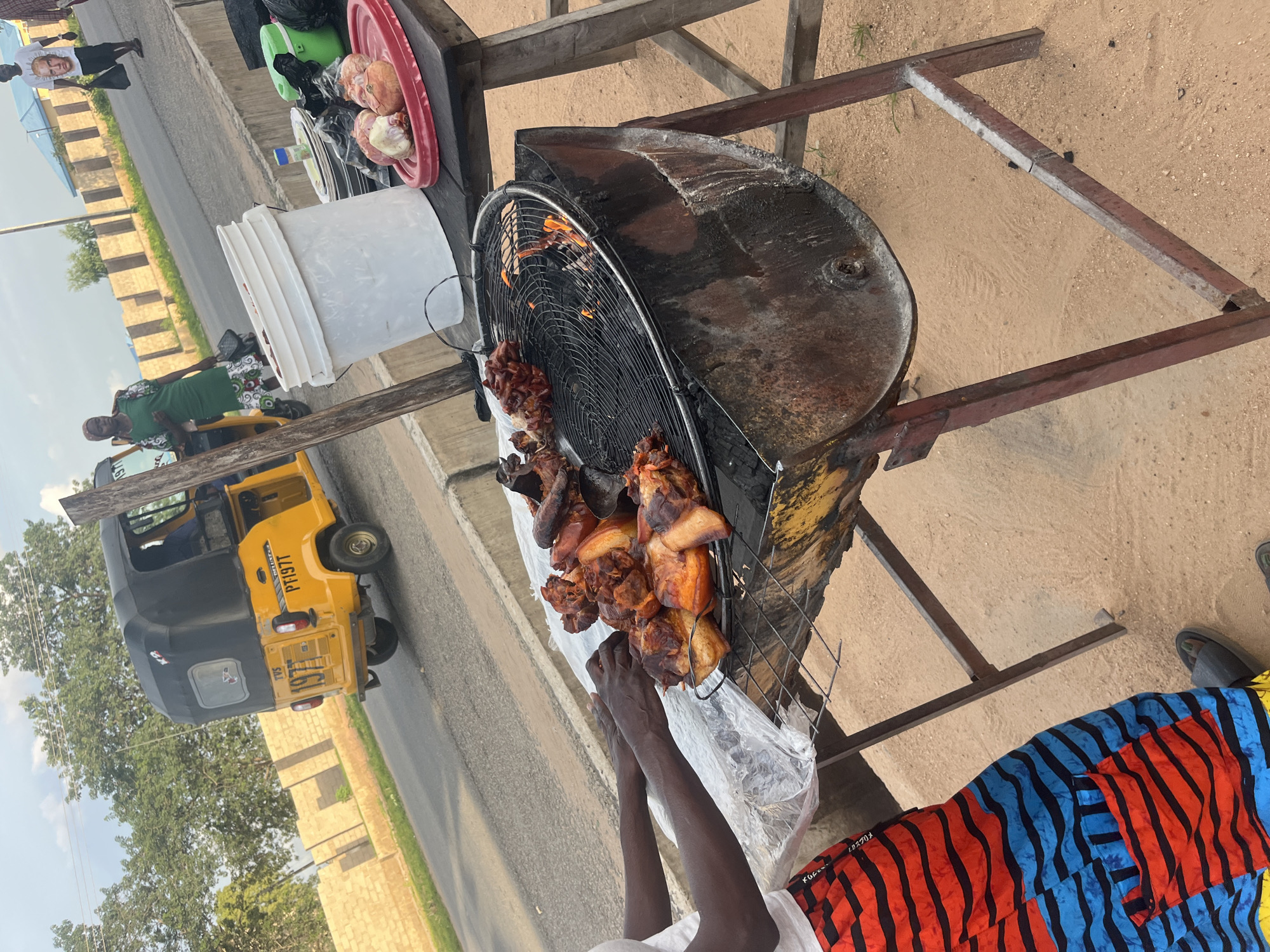 The image shows an outdoor scene in Jalingo, Nigeria, with a focus on a roadside food stall. The primary subject is a person grilling meat, likely chicken, over a makeshift grill fashioned from a barrel. The meat appears to be cooked to a golden brown and is arranged on a wire rack.
In the background, a yellow and black auto-rickshaw, commonly known as a "Keke," is visible, suggesting a busy street environment. Other objects at the stall include a red bowl containing what appear to be onions or garlic, a green cooler, and a white bucket. There are also glimpses of other people in the background, including a woman in a green top and a person wearing a shirt with an image on it.
The setting is a sandy roadside with a paved surface and a low wall or curb. Trees and buildings are visible in the distance. The lighting suggests it is daytime, possibly late morning or afternoon, with natural sunlight casting shadows. The overall impression is of a typical street-side business in a bustling Nigerian town.
Notable details include the rustic nature of the grilling equipment and the vibrant colors of a patterned fabric worn by someone in the foreground. There is some visible text on the auto-rickshaw, including the license plate "PT197T" and the brand name "TVS." The scene depicts the daily activity of a street vendor preparing and selling food.