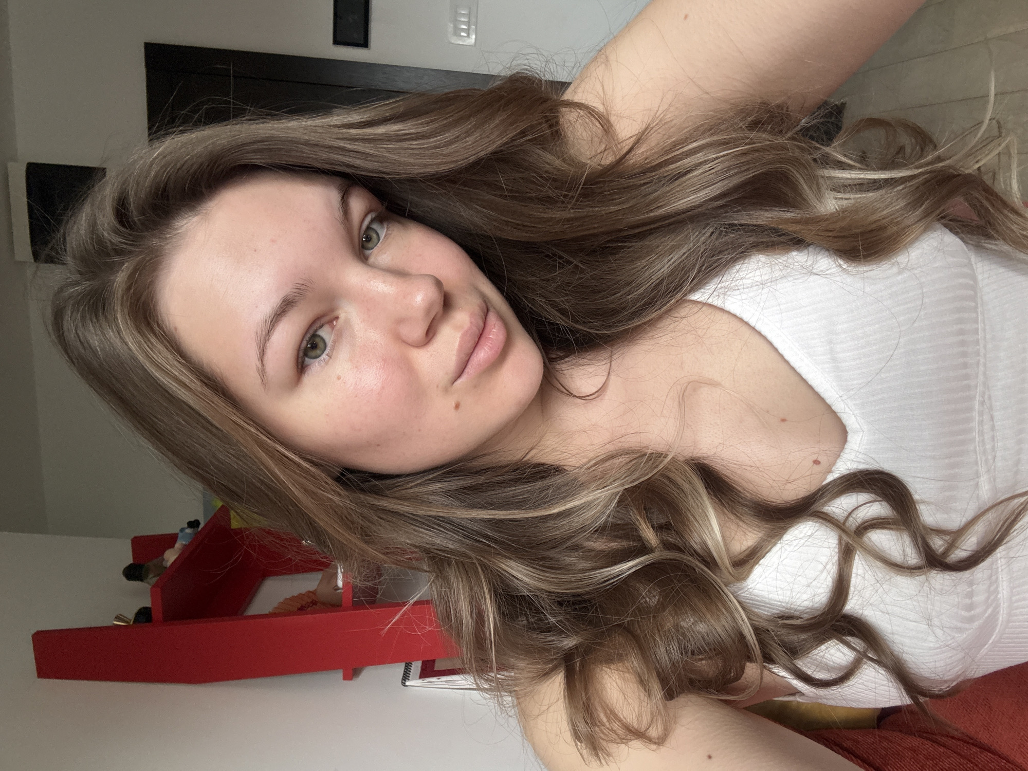 The image displays a close-up portrait of a young woman with long, wavy, light brown hair that has subtle blonde highlights. Her hair cascades around her head and shoulders. She has green eyes, fair skin, and a neutral expression with slightly parted lips, looking directly towards the camera. She is wearing a white, ribbed tank top or camisole.
The setting appears to be indoors. In the background, to the viewer's left, a bright red, multi-tiered shelf or organizer is visible, holding a few small, indistinct objects. To the woman's right and above, parts of a light-colored wall are seen, adorned with two dark, rectangular frames or decorative elements. The lighting is soft and even, typical of an indoor daytime setting.
The primary activity depicted is the woman posing for a photograph, likely a selfie, given the close perspective. Her demeanor is calm and natural. There are no overt location cues visible within the image to specify a geographical location, although the provided context indicates Vozdovac Urban Municipality, Serbia. No visible text is present in the image.
