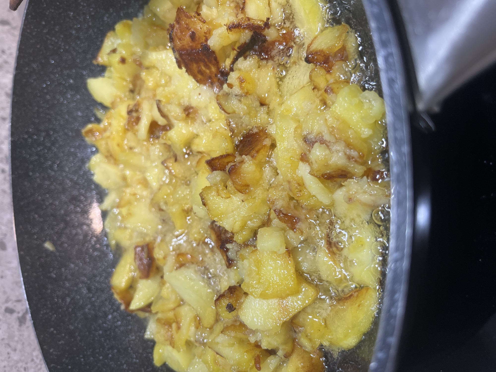 The image shows a close-up of fried potatoes being cooked in a pan. The potatoes are cut into irregular chunks and appear golden brown and crispy, with some pieces showing darker, more caramelized edges. They are submerged in hot oil, indicated by the bubbling around them. The pan itself is dark and appears to be non-stick, with a slight sheen. The background is blurred, but hints of a countertop with a speckled pattern are visible on the left, and a dark, smooth surface, likely a stovetop, is on the right. The lighting suggests an indoor setting, possibly a kitchen. The focus is entirely on the food and the cooking process, with no people or other objects prominently featured. The overall impression is one of active food preparation, with the focus on the visual appeal of the browning potatoes.