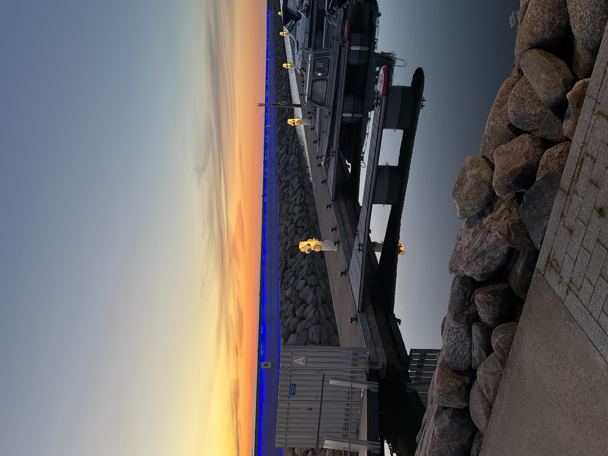 The image depicts a marina at sunset in Tallinn, Estonia. The sky displays a gradient of colors, from a pale blue at the top to vibrant orange and yellow hues near the horizon, with wispy clouds. A long, straight structure with blue lighting runs along the horizon in the distance, separated by a wall of large, gray rocks. In the foreground, a wooden pier extends into the dark water, with several boats moored alongside. The pier is illuminated by yellow bollard lights. On the left side of the pier, a utilitarian gray structure with a door and a sign bearing the letter "A" is visible. The water reflects the lights and the surrounding structures. The edge of the marina is lined with large, rough-hewn rocks and a paved walkway. There are no people visible in the image.