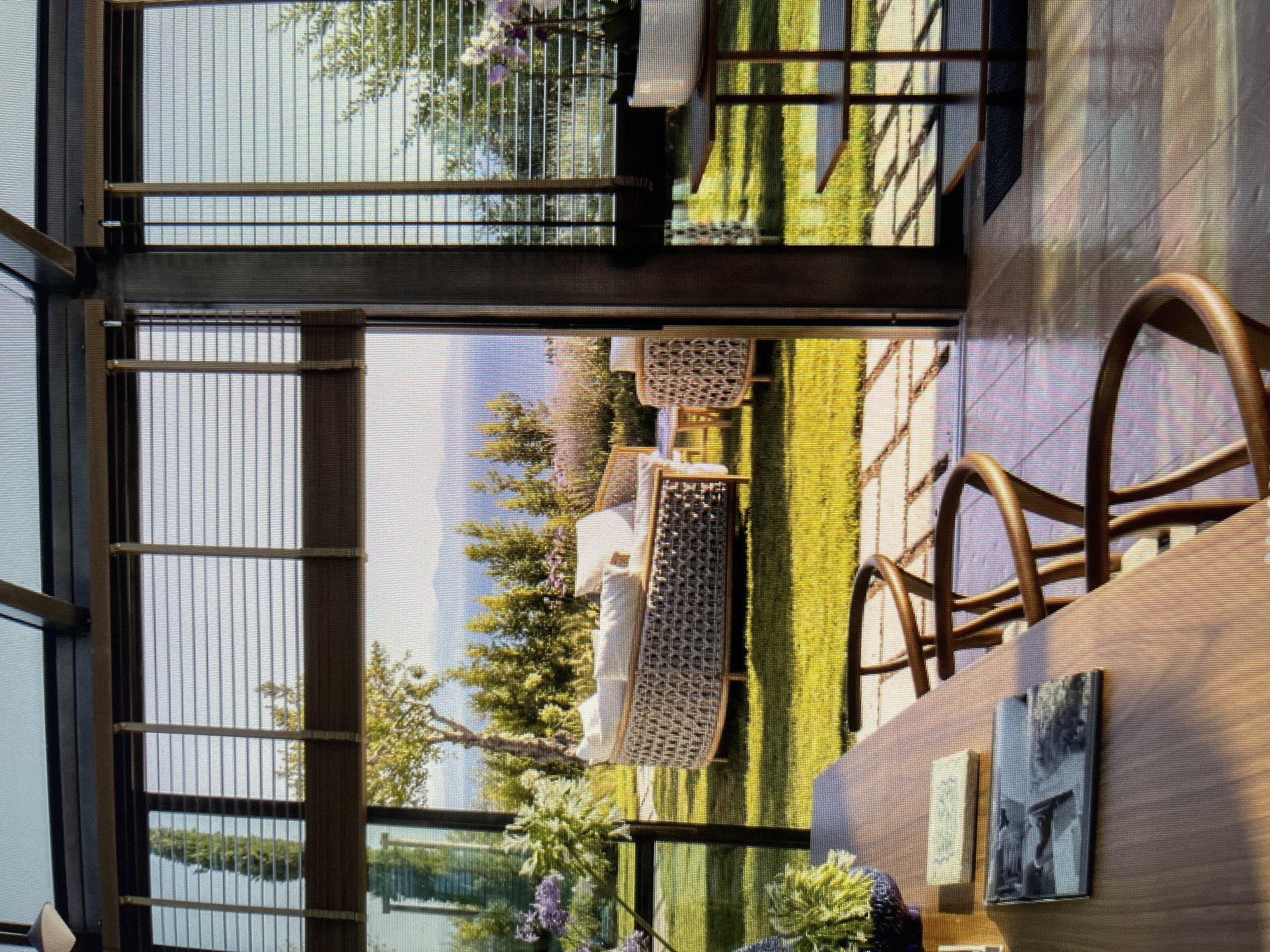 The image shows a sunlit outdoor seating area viewed from an indoor dining space. The indoor area features a wooden table with two books and a potted plant. Three wooden chairs with curved backs are visible around the table. Beyond the indoor space, large glass doors open to an exterior patio with stone pavers.
The exterior features a neatly manicured lawn with a round, woven outdoor sofa and chairs. In the background, a body of water is visible with hills beyond. The scene is framed by tall trees and lush greenery. The lighting suggests it is daytime, with strong sunlight creating shadows. The architectural elements, including the glass doors and roof structure, indicate a modern design. There are no people visible in the image. The location is not in Łódź, Poland.