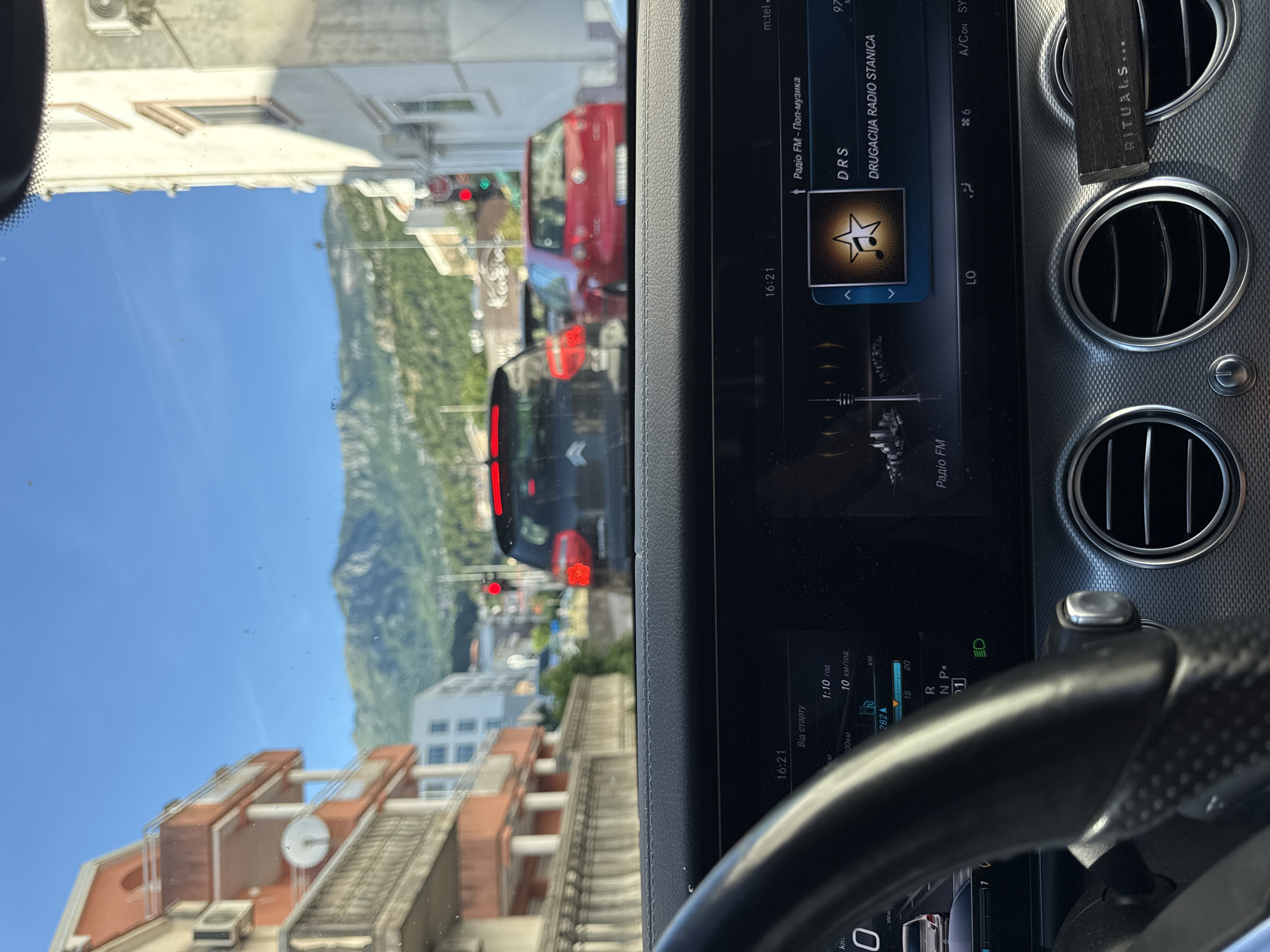 The image shows a view from inside a car, looking forward through the windshield. The car is stopped in traffic on a street in Budva, Montenegro. In the foreground, the dashboard of the car is visible, including the steering wheel and a digital display showing the time as 16:21, as well as radio information for "DRS DRUGACIJA RADIO STANICA". Two red brake lights are illuminated on the car in front. Other vehicles are visible in the traffic, including a black hatchback and a red hatchback. Buildings line the street on either side, and in the background, a mountain range is visible under a clear blue sky. The street appears to be a main thoroughfare with traffic lights and street signs.