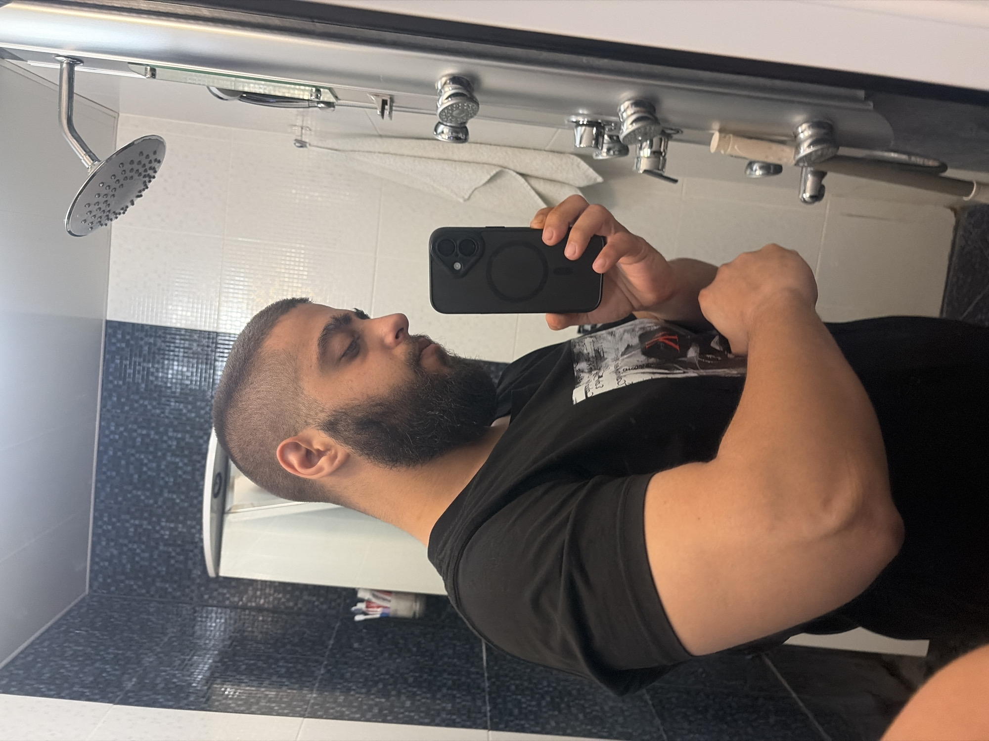 A man with a short haircut and a beard is taking a photo of himself in a bathroom using a black smartphone. He is wearing a black t-shirt. The bathroom has a showerhead, white and black tiled walls, and a white sink. A white towel is visible hanging above the sink.