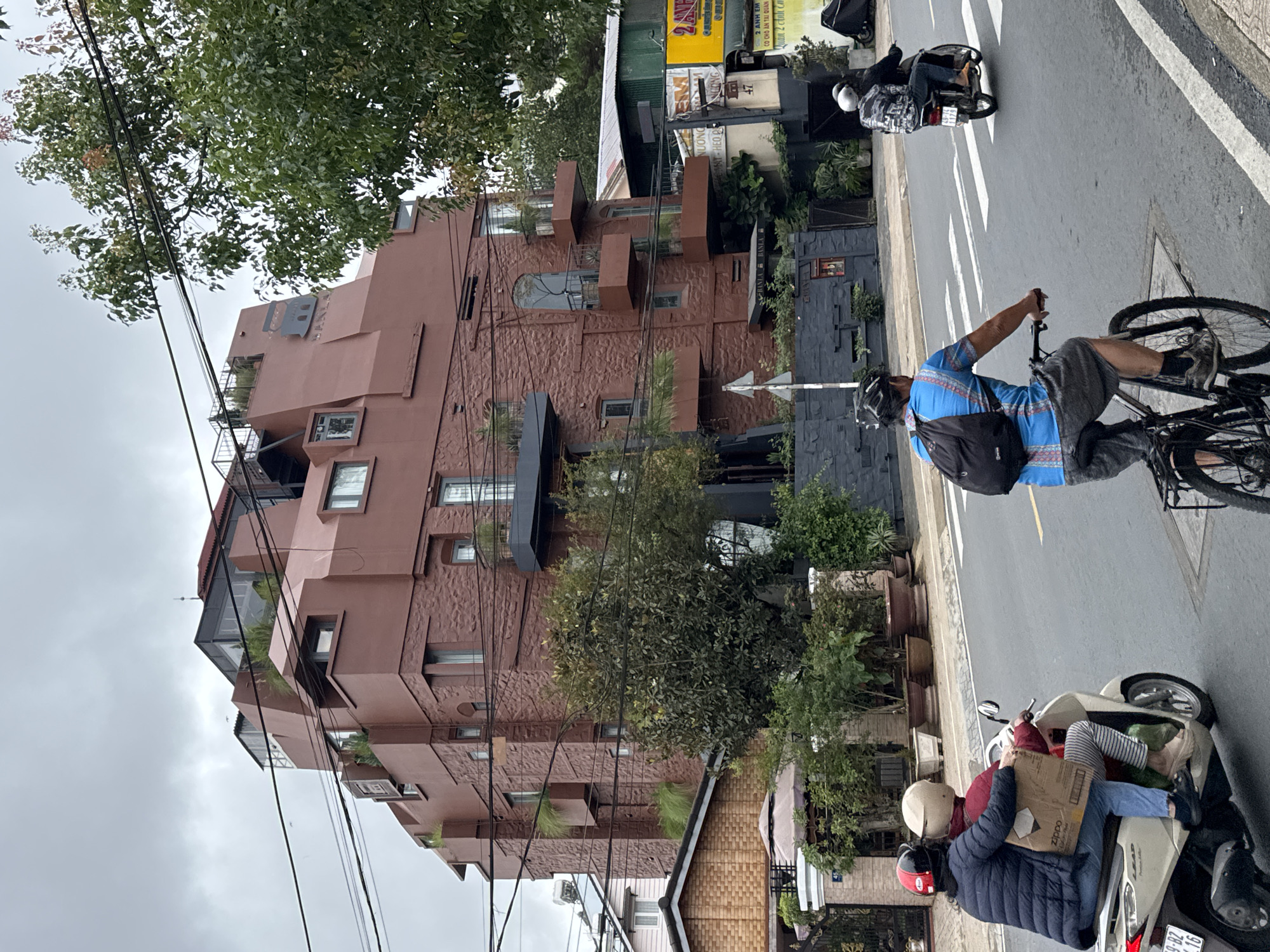 The image captures a street scene in Lâm Viên - Đà Lạt, Vietnam, on a cloudy day. The most prominent feature is a unique multi-story building with a brown, angular, and textured facade, possibly brick or stone, featuring irregularly placed windows and some visible greenery on balconies. A metallic "LB" sign is affixed to the upper section of this building.
On the street, several individuals are engaged in daily activities. A person wearing a blue patterned shirt, dark shorts, and a helmet is riding a bicycle, carrying a black backpack. Further down the road, another individual is seen on a motorbike with a sidecar. Closer to the foreground, two people are riding a scooter. The front rider, wearing a red jacket and beige helmet, holds a cardboard box with a "zippo" logo visible. The passenger, in a dark blue puffer jacket and red helmet, sits behind. The scooter's license plate, "49-HZ 01.16", is partially visible.
The paved road features white dashed lines and a yellow line. Overhead, numerous black power lines crisscross the frame, partially obscured by lush green tree foliage. In the background, across the street from the distinctive brown building, other structures are visible, including one with several Vietnamese signs. Text visible on these signs includes "2 ANH EM", "CO CHO ÂN TẠI QUAN", "Quan 2 Anh C", "NƯƠNG", and "MAT-HEO". The overcast sky suggests a subdued daylight.