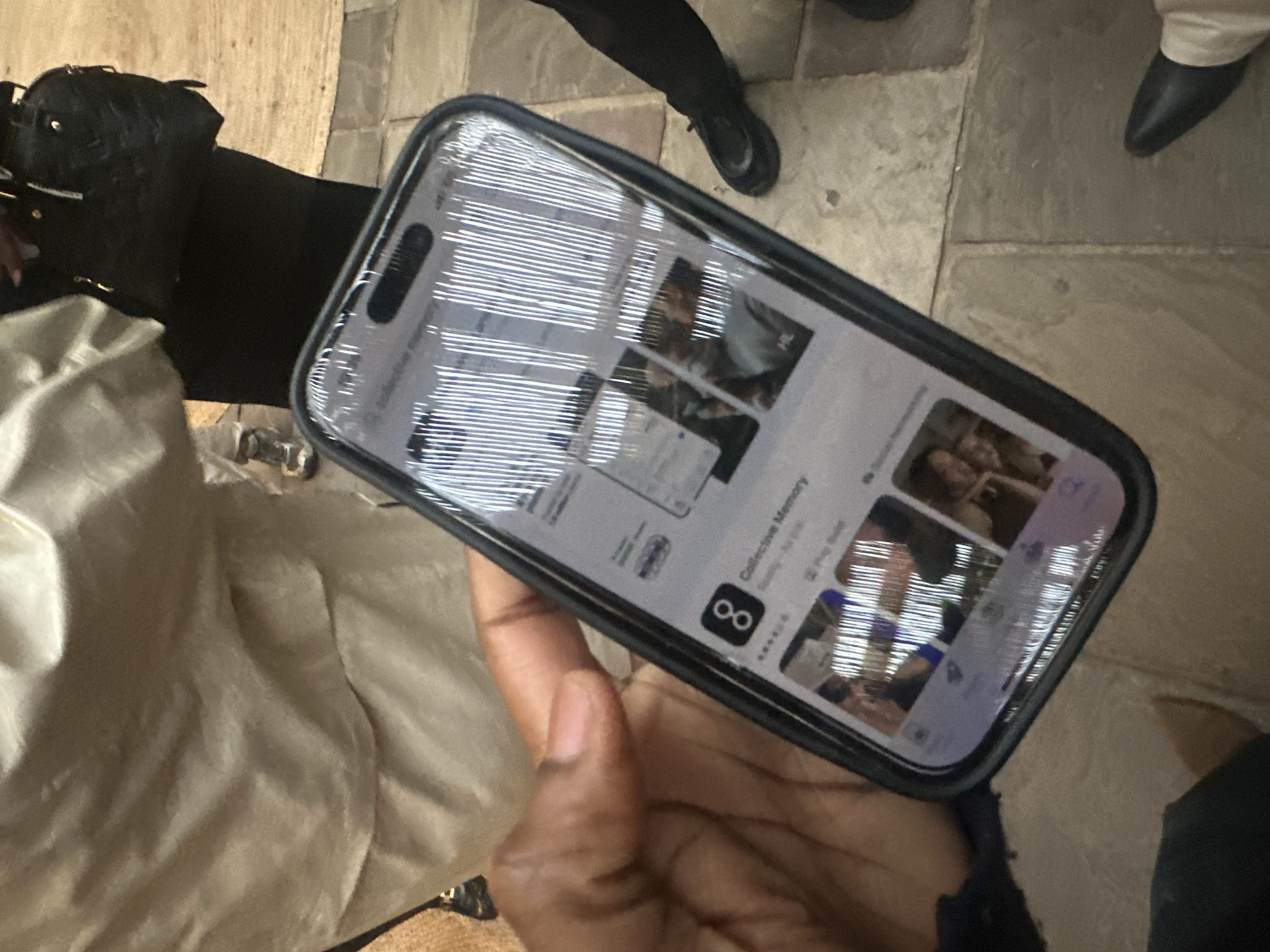 A medium shot captures a hand holding a phone displaying the App Store with the "Collective Memory" app. The background shows elements of urban attire: a black purse, beige fabric, black clothing, and various shoes, indicating an environment likely occupied by people. The flooring appears to be paved, suggesting an outdoor or public location in an urban setting. The reflection in the phone screen features what appears to be window blinds. The presence of the App Store and the urban attire imply a contemporary urban environment. No specific landmarks identify the location as London, United Kingdom.