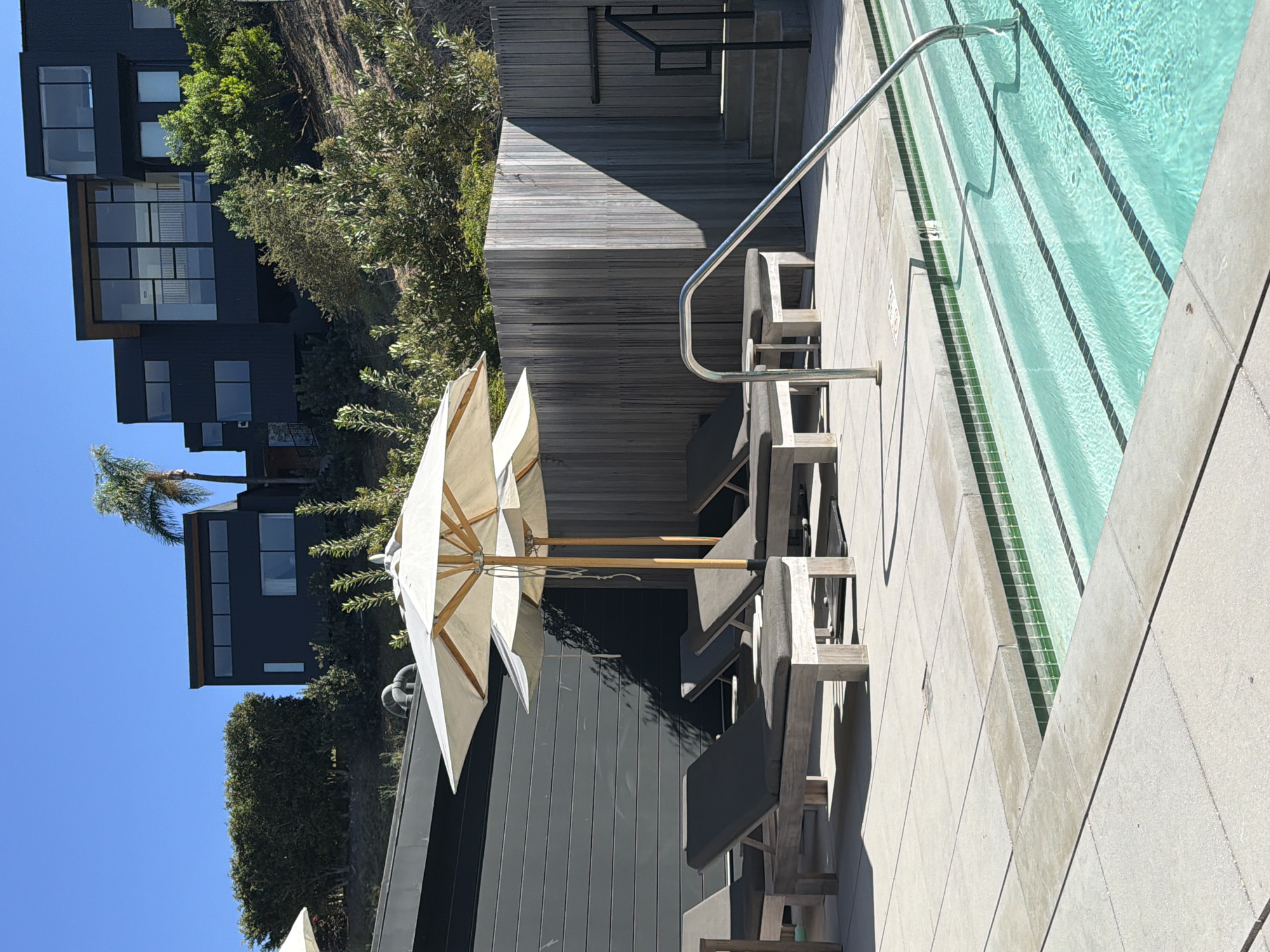 In Malibu, California, a modern, multi-story building with a dark facade and numerous windows is situated on a hillside covered with dense green foliage. In the foreground, a swimming pool with clear turquoise water and a stepped entry, lined with green tile, is visible. Beside the pool, several contemporary lounge chairs with dark cushions are arranged on a paved patio. Two large, light-colored umbrellas stand over the lounge chairs, providing shade. A rustic wooden fence borders the patio area, and a metal handrail curves into the pool. The sky is a clear, bright blue.