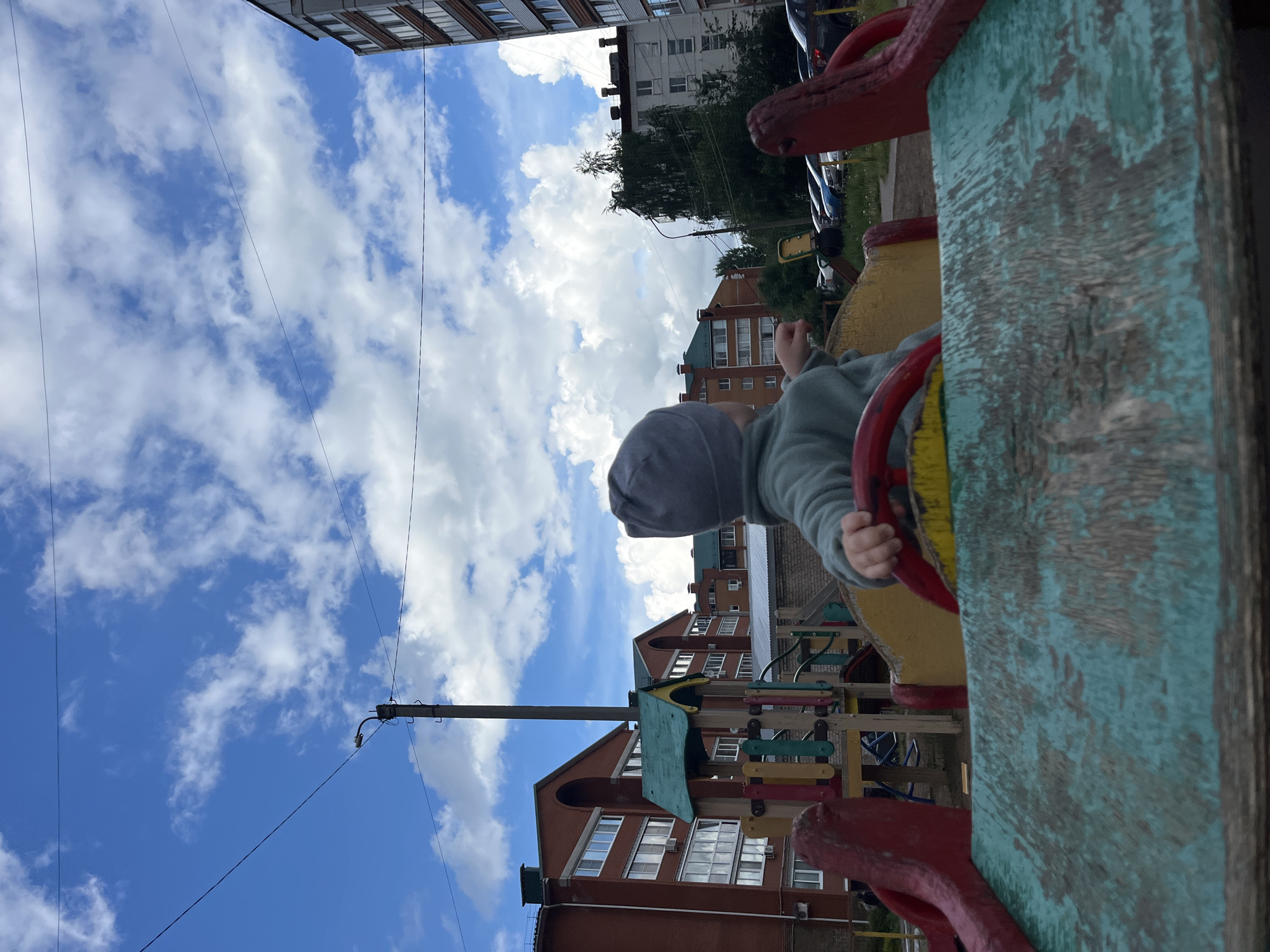 The image captures an outdoor scene at a playground, viewed from a low angle, looking upwards towards the sky and surrounding buildings. A young child, dressed in a grey hooded top and a light blue/grey long-sleeved garment, is visible from the back and right side, actively engaged with a wooden playground structure. Their right hand is grasping a red steering wheel-like handle.
The foreground is dominated by a weathered wooden play structure, possibly designed as a car or train, featuring a prominent teal or blue-green painted surface with significant wear, revealing the wood beneath. Red painted elements, including the handles the child is holding and other decorative parts, are also visible. To the left of the child, another section of the playground structure can be seen with colorful horizontal bars in red, yellow, blue, and green.
In the mid-ground and background, the scene is framed by multi-story residential buildings, consistent with apartment blocks. Some buildings are constructed from red brick, while others have lighter colored facades. Trees with green foliage and parked cars are visible between the buildings and the playground area. A utility pole or lamppost stands prominently, extending towards the sky.
The weather appears bright and clear, with a vivid blue sky adorned with numerous fluffy white cumulus clouds. The time of day is daytime, likely late morning or afternoon, given the bright illumination and cloud cover. No discernible text is visible within the image. The setting suggests a typical urban or suburban playground in Chaikovsky, Russia.