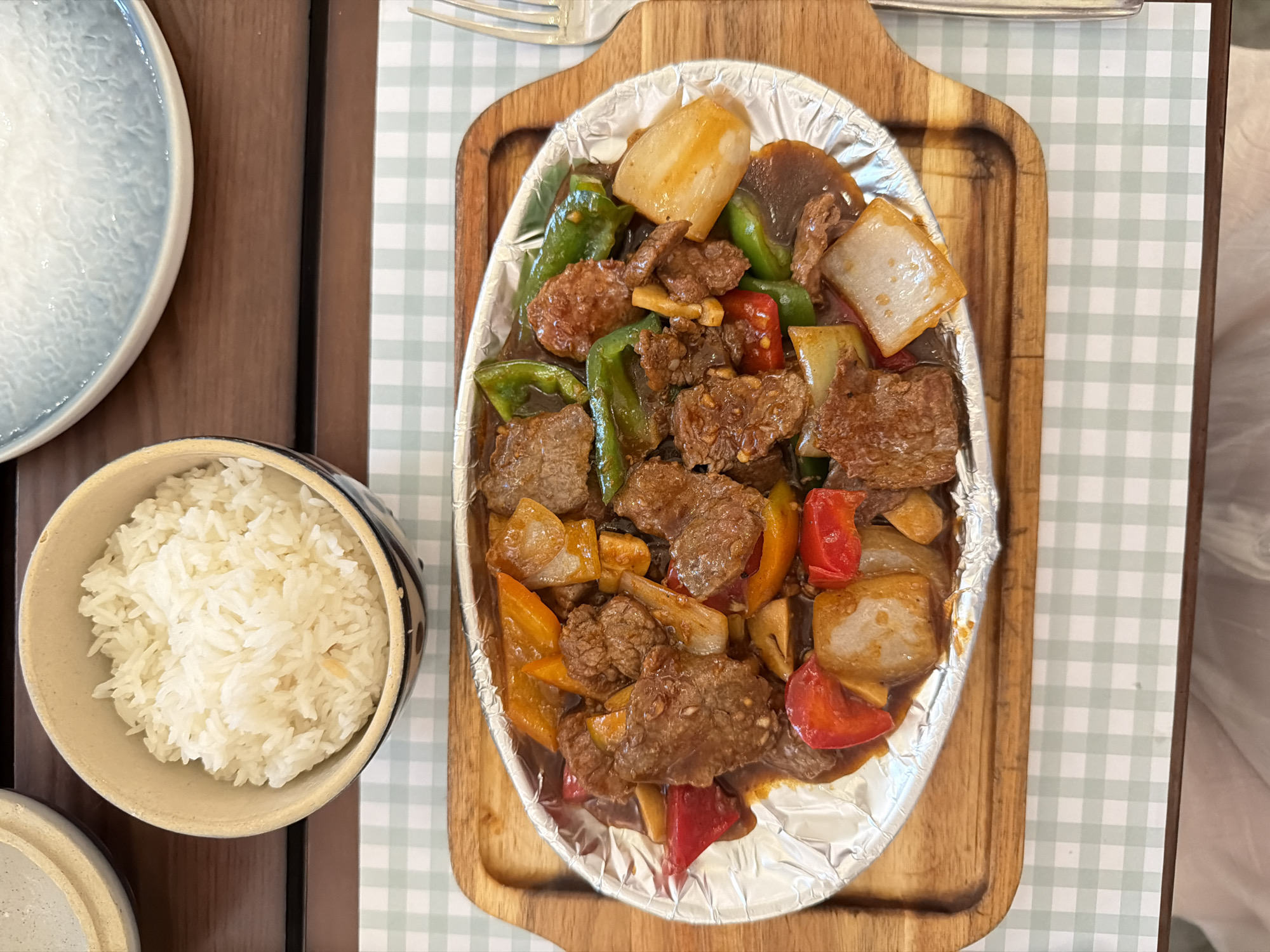 The scene is a tabletop dining setup located in Nha Trang Ward, Vietnam. A main dish is presented on an oval metal platter lined with crinkled aluminum foil. This platter contains a stir-fried dish featuring numerous dark brown, irregular chunks of meat (consistent with beef), mixed with large pieces of vibrant green, red, and orange bell peppers, as well as white-translucent pieces resembling onions or possibly water chestnuts. All ingredients are coated in a glossy, dark brown sauce. This platter rests on a light brown wooden serving tray with subtly raised edges.
To the upper left of the wooden tray, a light brown ceramic bowl holds a generous portion of white, long-grain rice. Partially visible in the upper right background is the rim of a light blue/grey ceramic plate with a textured, slightly uneven surface. A silver-colored metal fork with four tines is positioned on the right side of the wooden serving tray.
The entire arrangement is situated on a light green and white checkered placemat. The underlying surface is a dark brown wooden table. In the lower right corner of the frame, a segment of light-colored clothing and a person's forearm are partially visible, indicating the presence of a diner.
