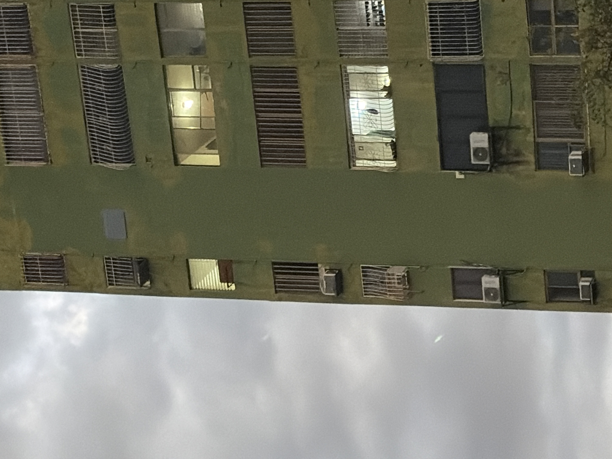 The image shows the facade of a multi-story building in Ciudad Guayana, Venezuela, against a cloudy sky. The building has a greenish exterior and features a series of windows, some with security bars or grilles. Some windows appear to be illuminated from within. Several air conditioning units are affixed to the exterior of the building. No people are visible in the image.