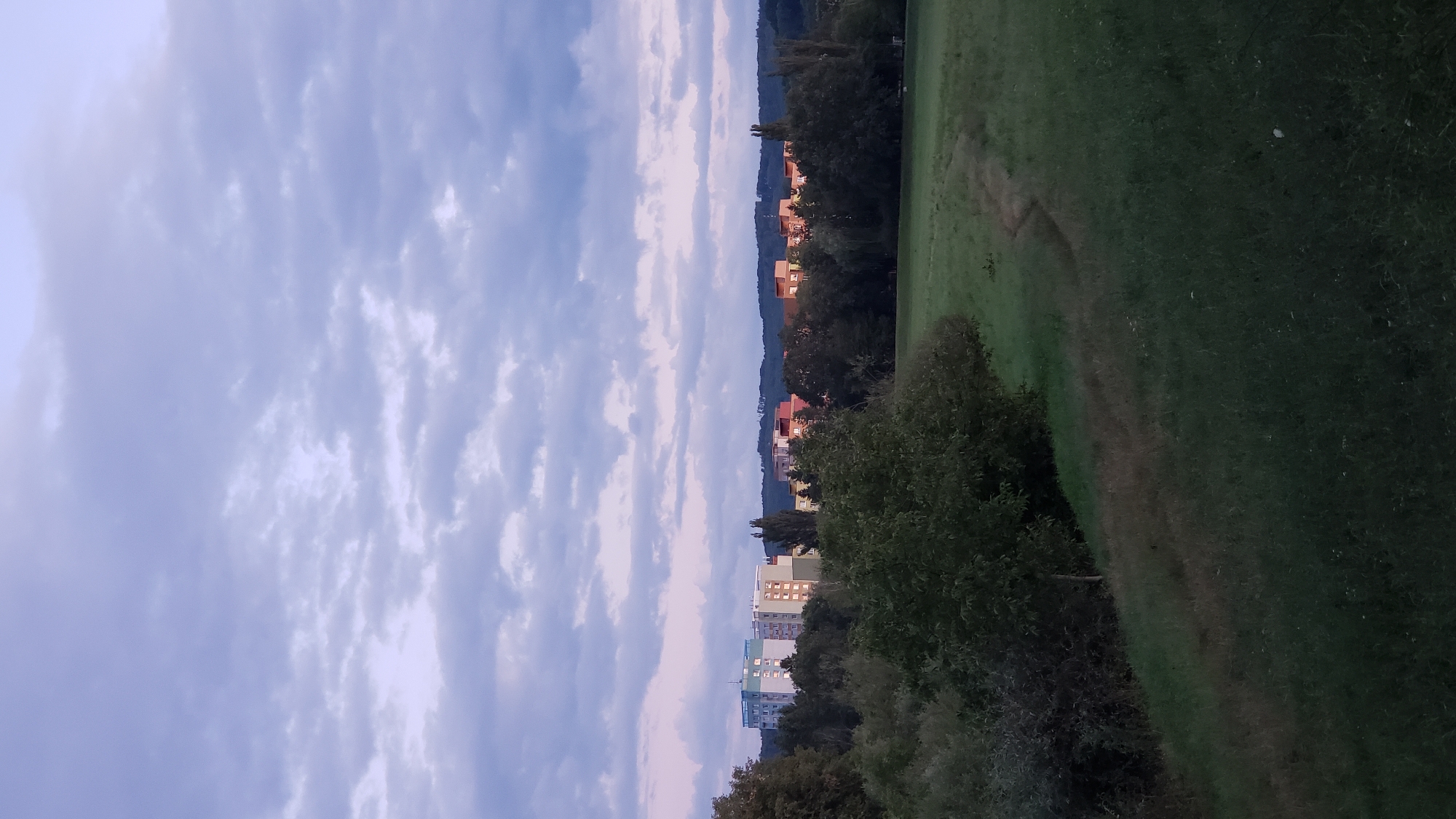 The image depicts a panoramic view of the city of Prešov, Slovakia, at dusk. In the background, several apartment buildings of varying heights and colors are visible, nestled among a line of trees. The buildings are illuminated by artificial lights, casting a warm glow against the dusky sky. The sky itself is filled with dramatic clouds, tinged with shades of purple and pink, suggesting the last moments of daylight.
In the foreground, a vast expanse of green grass slopes gently downwards. A narrow, winding path is visible cutting through the grass, its darker color indicating it is well-trodden. To the left, dense foliage, including trees and bushes, forms a dark, natural border to the open field. The overall impression is one of a peaceful, urban landscape transitioning into night, with a blend of man-made structures and natural elements.