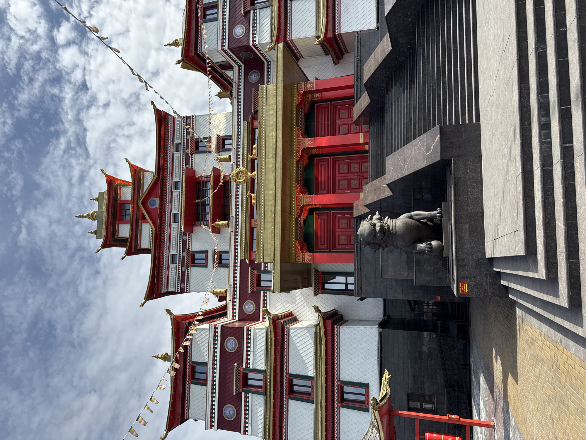 The image depicts a tall, multi-tiered Buddhist temple in Kyzyl, Russia, characterized by a mix of white, red, and gold architectural elements. The temple's facade is adorned with windows and decorative patterns. To the right of the temple is a dark gray, monolithic structure with steps leading up to it, and a lion statue at its base. The sky is visible in the background, with white clouds and colorful prayer flags strung between the temple structures. The ground near the bottom right of the frame is paved with bricks.