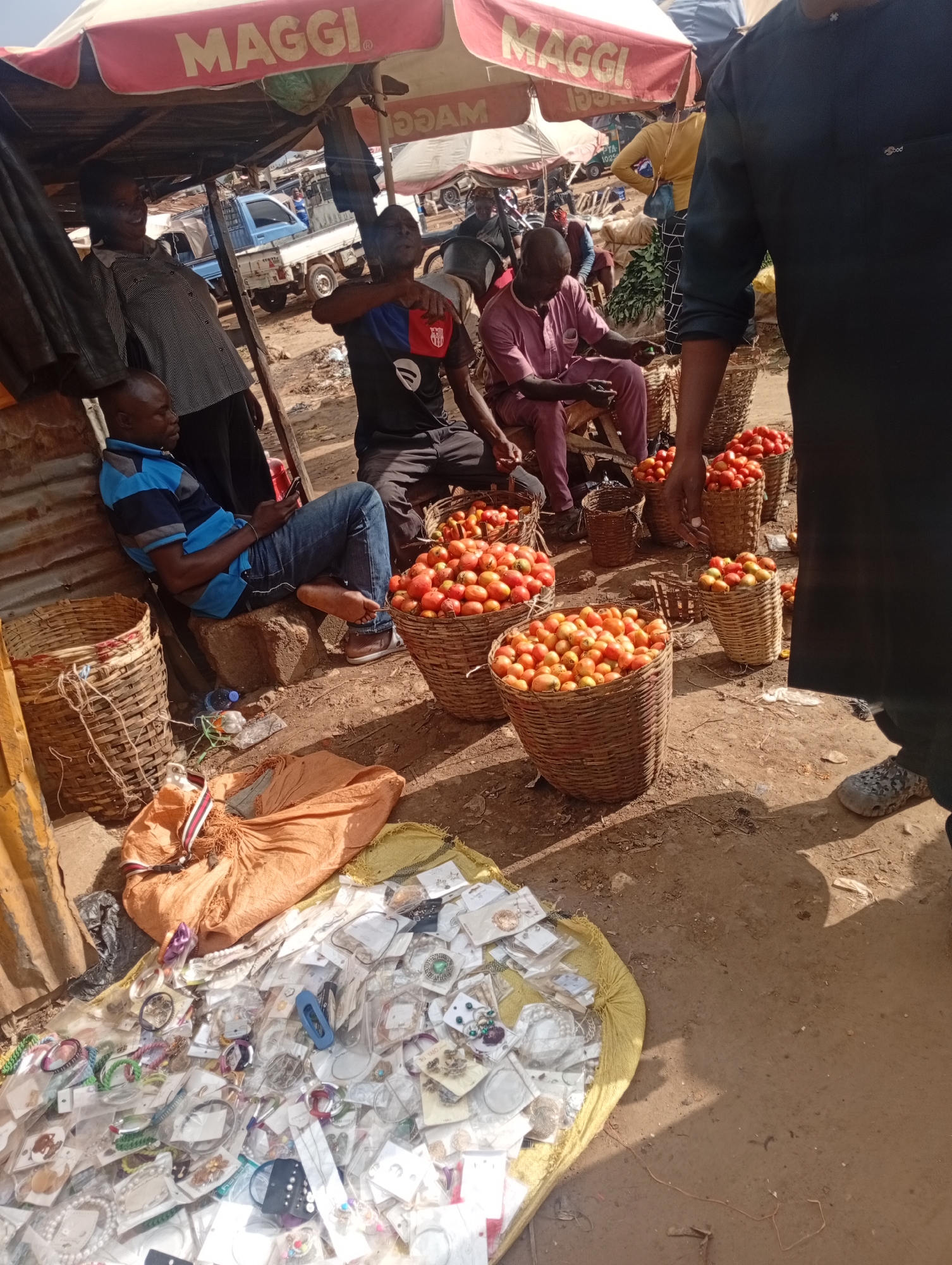 The image depicts an outdoor market scene in Rafin Par, Nigeria. Several individuals are present, engaged in various activities. In the foreground, a pile of assorted jewelry, including bracelets, necklaces, and earrings, is displayed on a yellow fabric, suggesting a retail transaction. To the left, a man wearing a blue and black striped shirt and jeans is seated, looking at his phone. He is positioned next to a woven basket.
Further into the scene, several large baskets filled with ripe tomatoes are prominently displayed, indicating produce sales. Men are seen tending to the produce or interacting with potential customers. One man in a pinkish-purple outfit is seated and holding a small green object, possibly a piece of fruit or a tool. Another man in a blue and red shirt is standing. Overhead, a red and white striped awning with the "MAGGI" logo is visible, providing shade and advertising. In the background, a light blue truck is parked, and other market stalls and people are faintly discernible.
The lighting suggests it is daytime, likely morning or afternoon, with bright sunlight casting shadows. The ground is dusty and uneven, characteristic of an outdoor market. The overall atmosphere appears to be one of commercial activity and daily life in a Nigerian marketplace. The people's expressions are difficult to discern clearly, but the scene conveys a sense of busyness and trade. The "MAGGI" text on the awning is a notable detail.