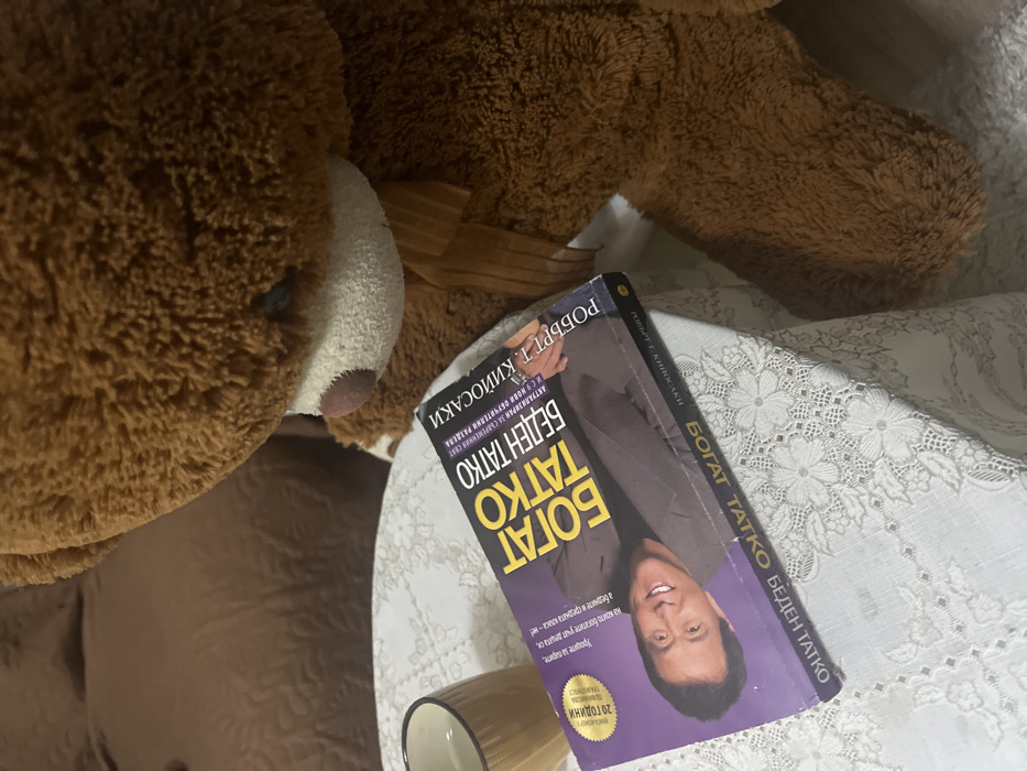 This image captures an indoor scene in Sedlartsi, Bulgaria, featuring a large brown teddy bear, a book, and a mug on a table. The teddy bear, positioned on the right, is a significant portion of the frame, displaying its head and upper torso. It has a light-colored snout, dark eyes, and a brown ribbon tied around its neck. The teddy bear appears to be leaning over or looking towards the table. On a white tablecloth with a floral lace pattern, a book titled "Богат Татко, Беден Татко" (Rich Dad Poor Dad) by Robert T. Kiyosaki is laid flat. The book cover features a smiling man. To the left of the book, a light brown or golden ceramic mug rests on the tablecloth. In the background, a brown upholstered surface, possibly a sofa or chair with a quilted pattern, is visible. No human individuals are present, and no active interactions are depicted beyond the static arrangement of objects.