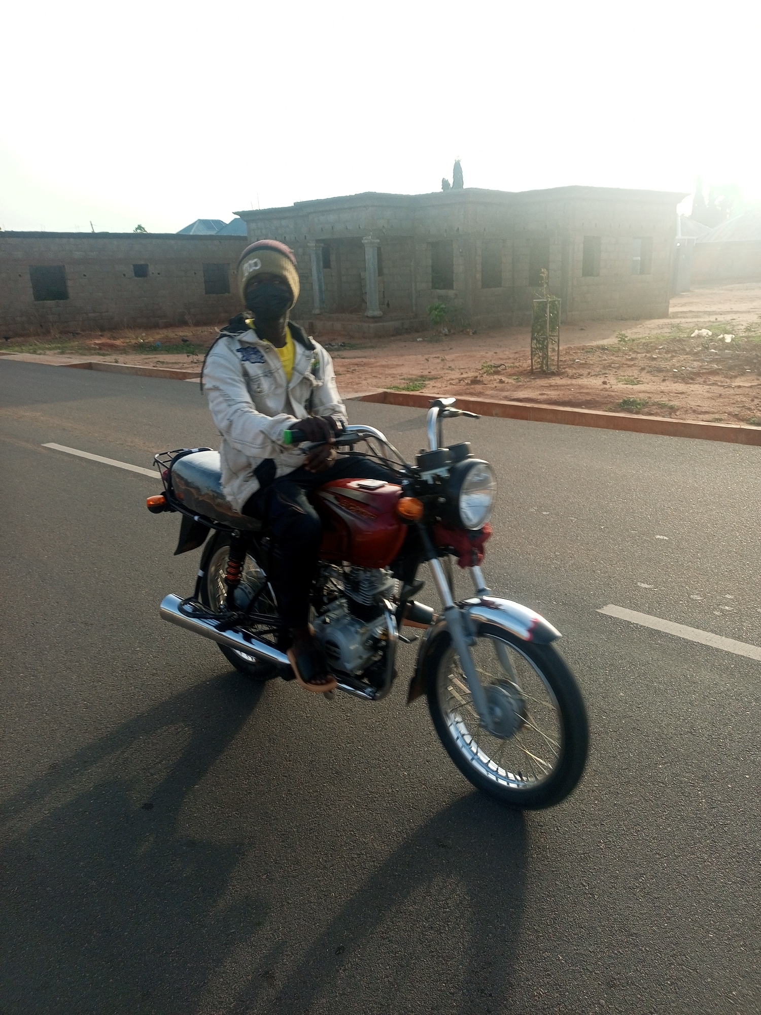 A person wearing a hat, face mask, white jacket, and black pants is riding a red motorcycle on an asphalt road in Tempure, Nigeria. The road has a white line marking. The background shows buildings under construction and a clear sky. The lighting suggests it is either early morning or late afternoon.