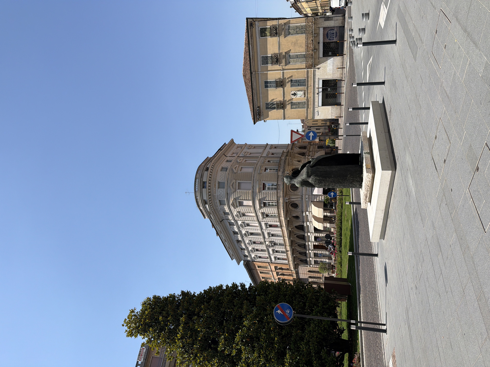 This photograph depicts a city square in Mantua, Italy. The angle of the shot is low, emphasizing the architectural height of the buildings that flank the square. In the foreground, a tree provides a natural border, obscuring the lower part of a road sign that prohibits pedestrians. A statue stands centrally, upon a plinth, and serves as a focal point. The square is paved with gray stone.
The buildings display classical Italian architecture with decorative facades. The architectural details include window frames, cornices, and balconies. There are figures on the balconies of one of the buildings.
The sky above is clear and bright blue.