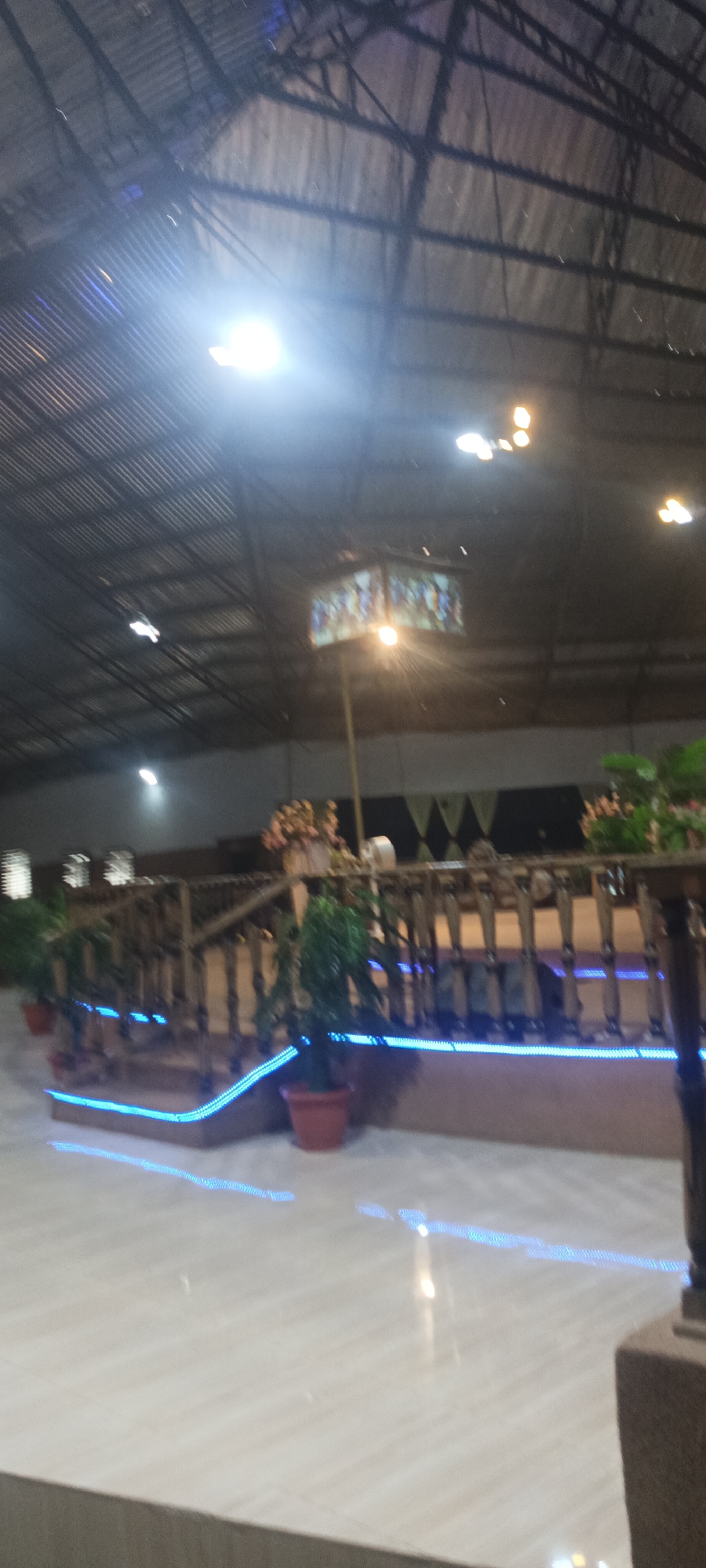 The image captures an indoor scene, possibly a venue or hall, with a corrugated metal roof supported by dark metal beams. A bright light illuminates the upper left portion of the frame, casting a glare. Below, a wooden railing with vertical balusters is decorated with blue LED strip lights, which are also present along the base of a raised platform. Several potted plants, including two green leafy plants in terracotta pots, are placed near the railing. In the background, decorative banners with a diamond pattern are visible, along with floral arrangements. The floor appears to be tiled and is reflecting the blue lights. The overall impression is of an illuminated event space with decorative elements. The location is specified as Rukuba Road, Nigeria.