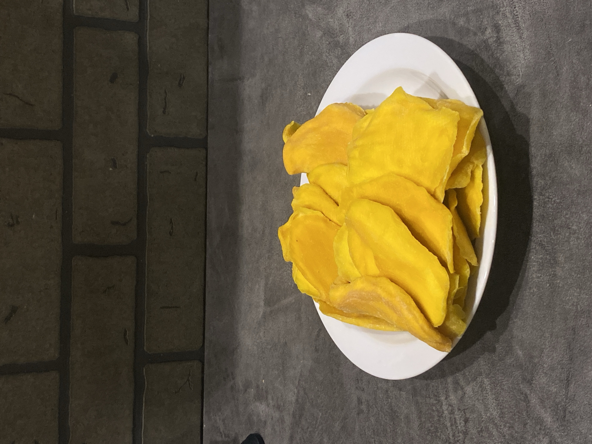 The image shows a white plate piled with dried mango slices. The plate sits on a dark, textured surface, possibly a countertop or table. To the left, a wall with a brick-like pattern can be seen. There are no people visible in the image. The location, Sochi, Russia, cannot be determined from the image.