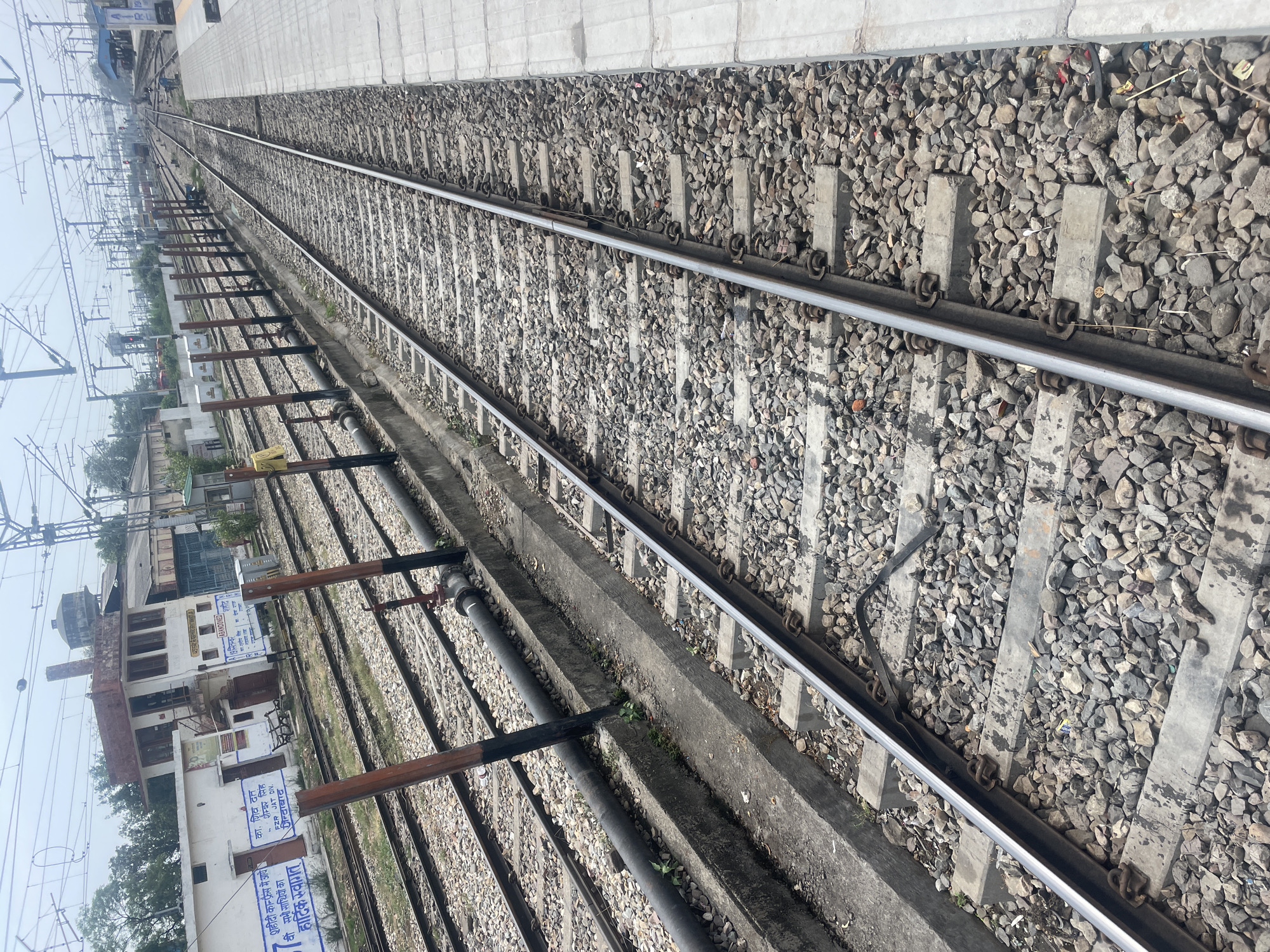 This is a photograph of a railway station in Pathankot, India. The image shows multiple sets of train tracks, ballast, and concrete sleepers. In the background, there is a small building with a red-tiled roof and a water tower. There are also overhead electrical wires for the trains. The scene is outdoors during the day, with a cloudy sky. The perspective is from an elevated position, looking down at the tracks. There are no people visible in the image.