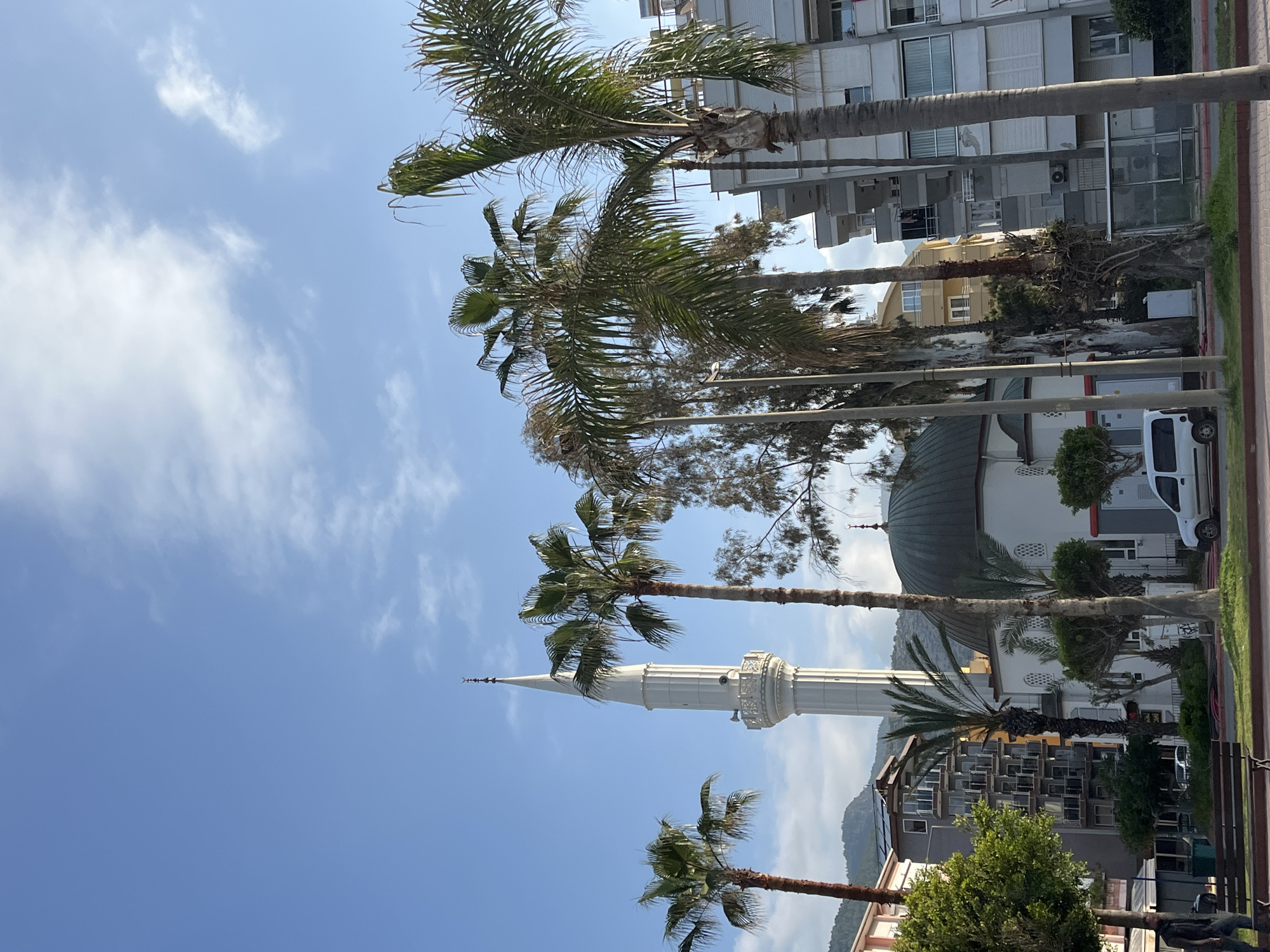 The image shows a street scene in Alanya, Turkey. The view is framed by palm trees on both sides, and the sky is blue with scattered clouds. In the background, a mosque with a tall, slender minaret and a green dome is visible. Several buildings are visible behind the mosque. A white van is parked on the side of the road near the mosque. The photograph appears to have been taken during the daytime, under bright sunlight.