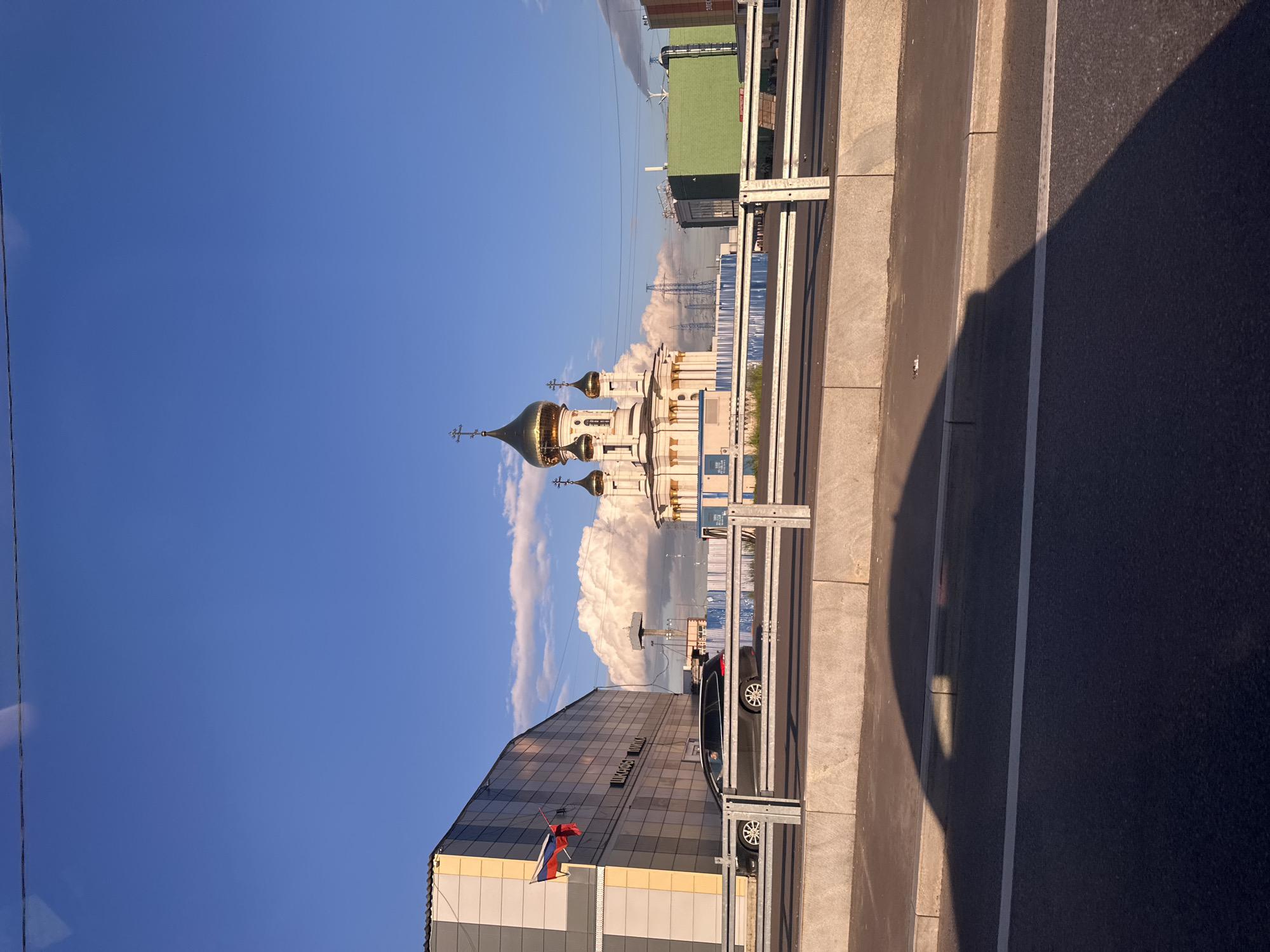 The image is a vertical, wide-angle shot of an urban landscape under a clear blue sky. In the foreground, a modern building with a reflective, glass-paneled facade is visible. A Russian flag flies from its side. Next to the building, a black car is parked, and a metal railing separates the sidewalk from a road.
In the middle ground, a golden-domed Orthodox church with multiple spires is prominent, its architecture contrasting with the modern buildings behind it. Large, white clouds are scattered across the sky, some appearing dense and puffy, suggesting a partly cloudy day. Further in the background, more modern buildings, some with green roofs, and power lines are visible, indicating a developed urban environment.
The lighting suggests it is late afternoon or early evening, with long shadows cast on the road and sidewalk, creating a dramatic contrast. The overall mood is serene and perhaps a little grand, with the golden domes of the church catching the sunlight. The scene appears to be set in a city, though the specific location is not identifiable from the image itself. There is some text visible on the reflective building which appears to read "ДИСКОНТ МИР".