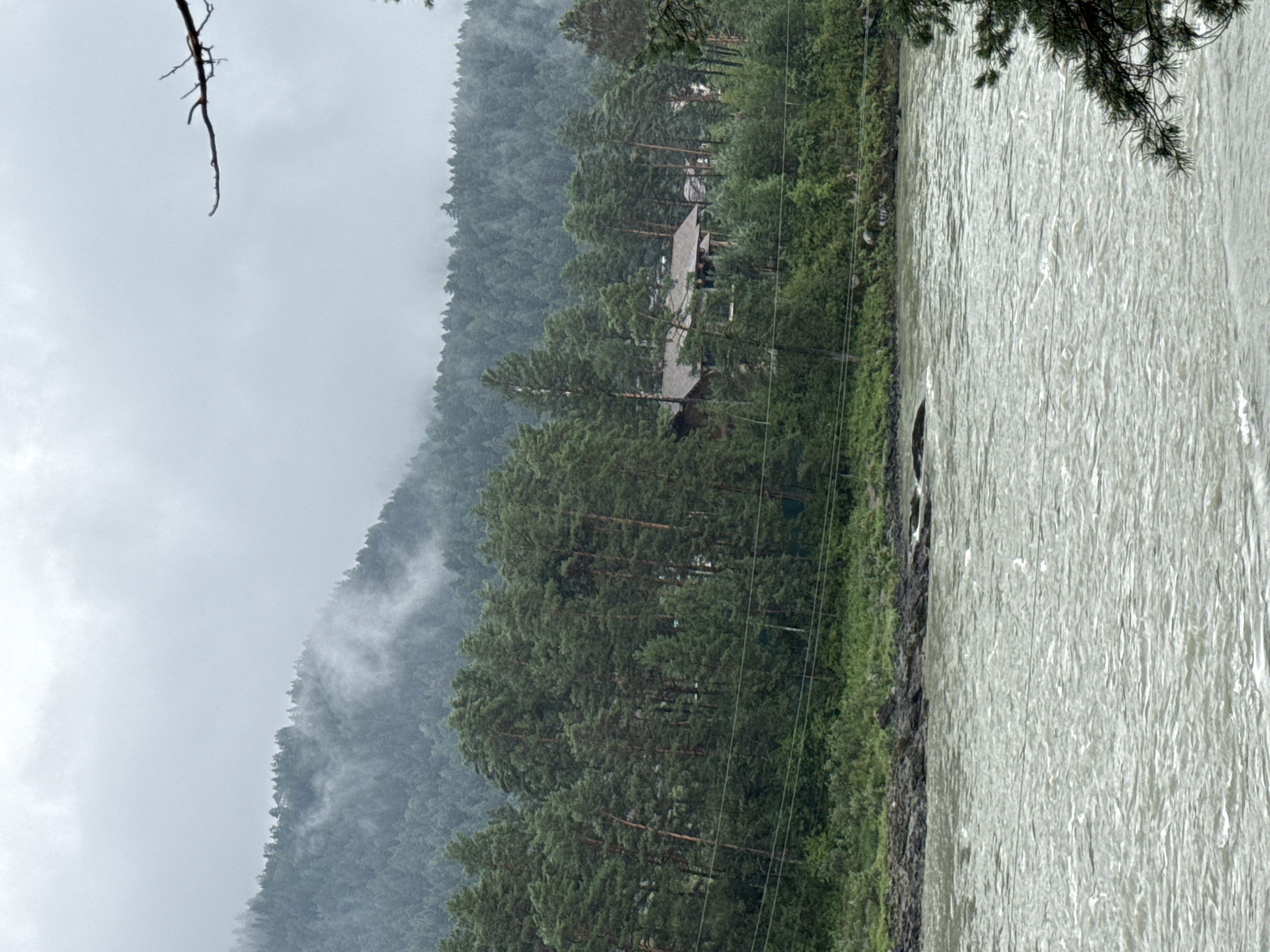 The image depicts a panoramic view of a river and a forested mountainside under an overcast sky. The foreground is dominated by a wide, choppy river with visible currents and some rocks along the shoreline. The middle ground features a dense forest of pine trees, with a cluster of buildings, possibly houses or lodges, nestled amongst them. The background shows a steep, mist-covered mountain slope covered in evergreen trees. The overall atmosphere is subdued and natural, suggesting a remote or natural environment. The location is identified as being in the city of Barangol, Russia. There are no people or active interactions visible in the image.