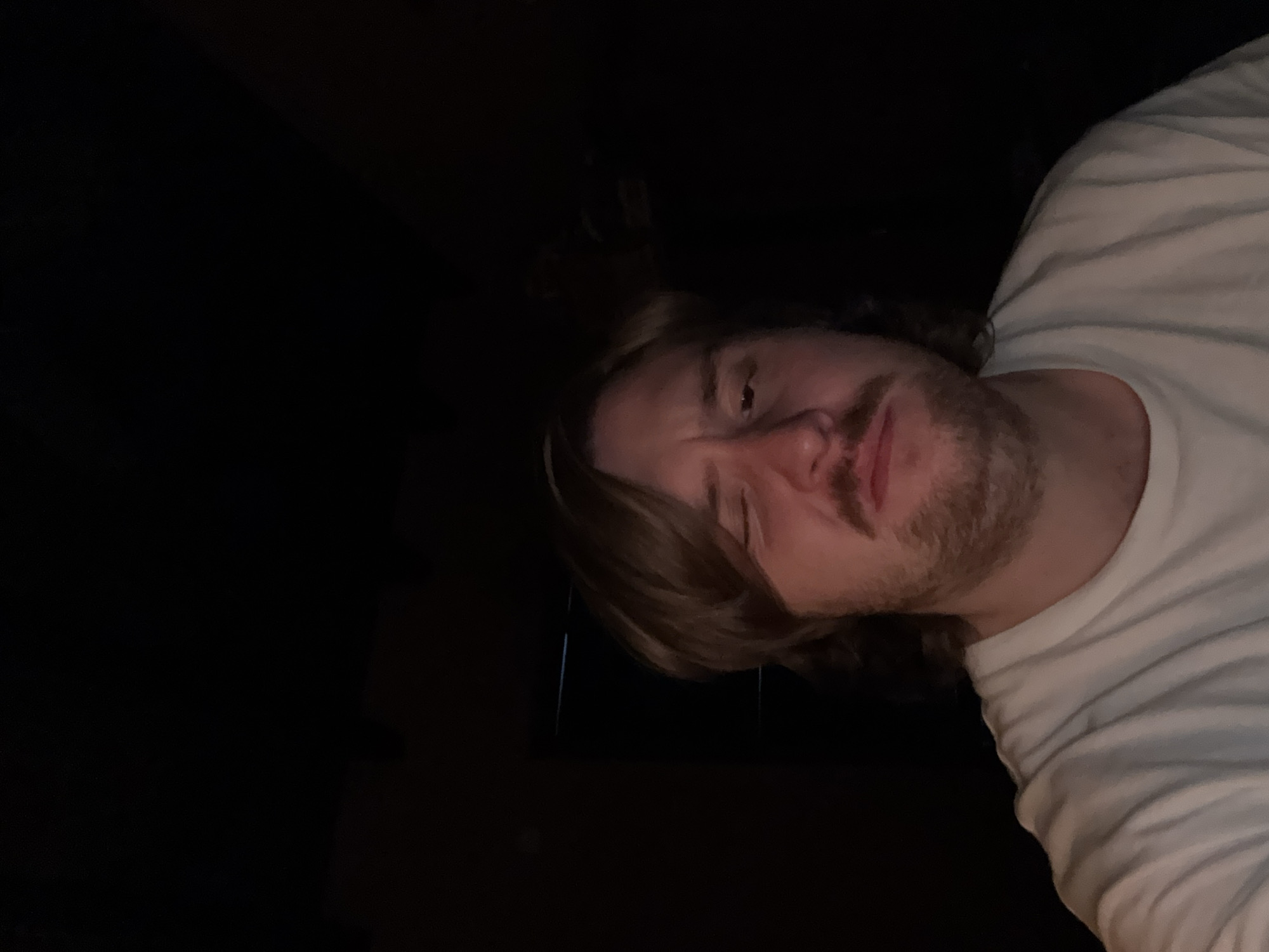 A man with light brown hair and a beard is visible from the chest up, wearing a white t-shirt. The background is mostly dark, with some indiscernible features. The photograph appears to be taken from a low angle, looking up at the man. No identifiable elements link the image to Cañada de Madero, Mexico.