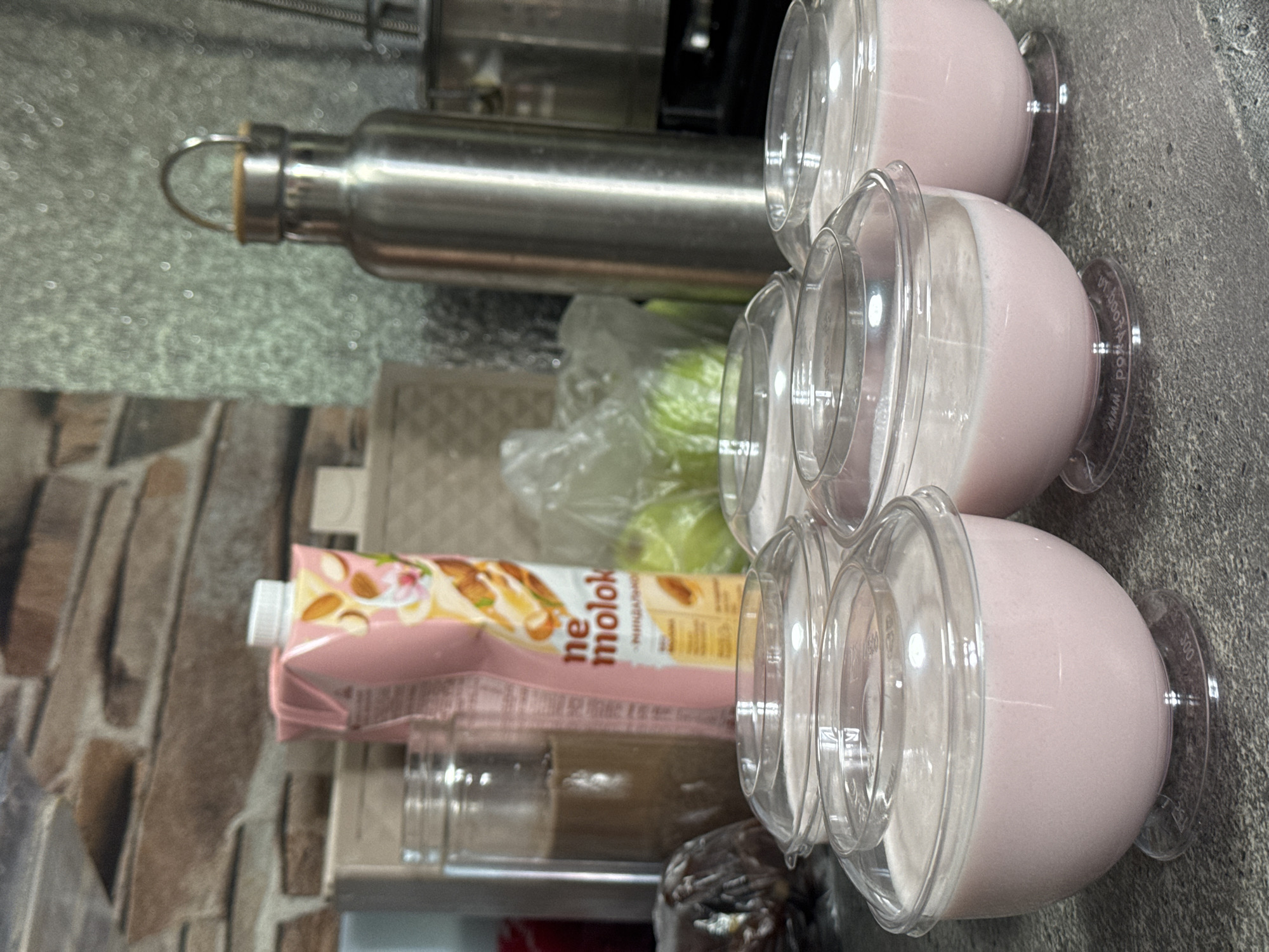 The image depicts a counter or shelf in what appears to be a kitchen or food preparation area. There are several items present: a stainless-steel water bottle, a carton of "ne molok" almond milk, possibly green apples in a clear plastic bag, some jars/containers, and four clear plastic cups filled with a pink liquid, possibly a yogurt or smoothie. The background includes a brick-like patterned wall. It is not possible to verify the location in Kurganinsk, Russia.