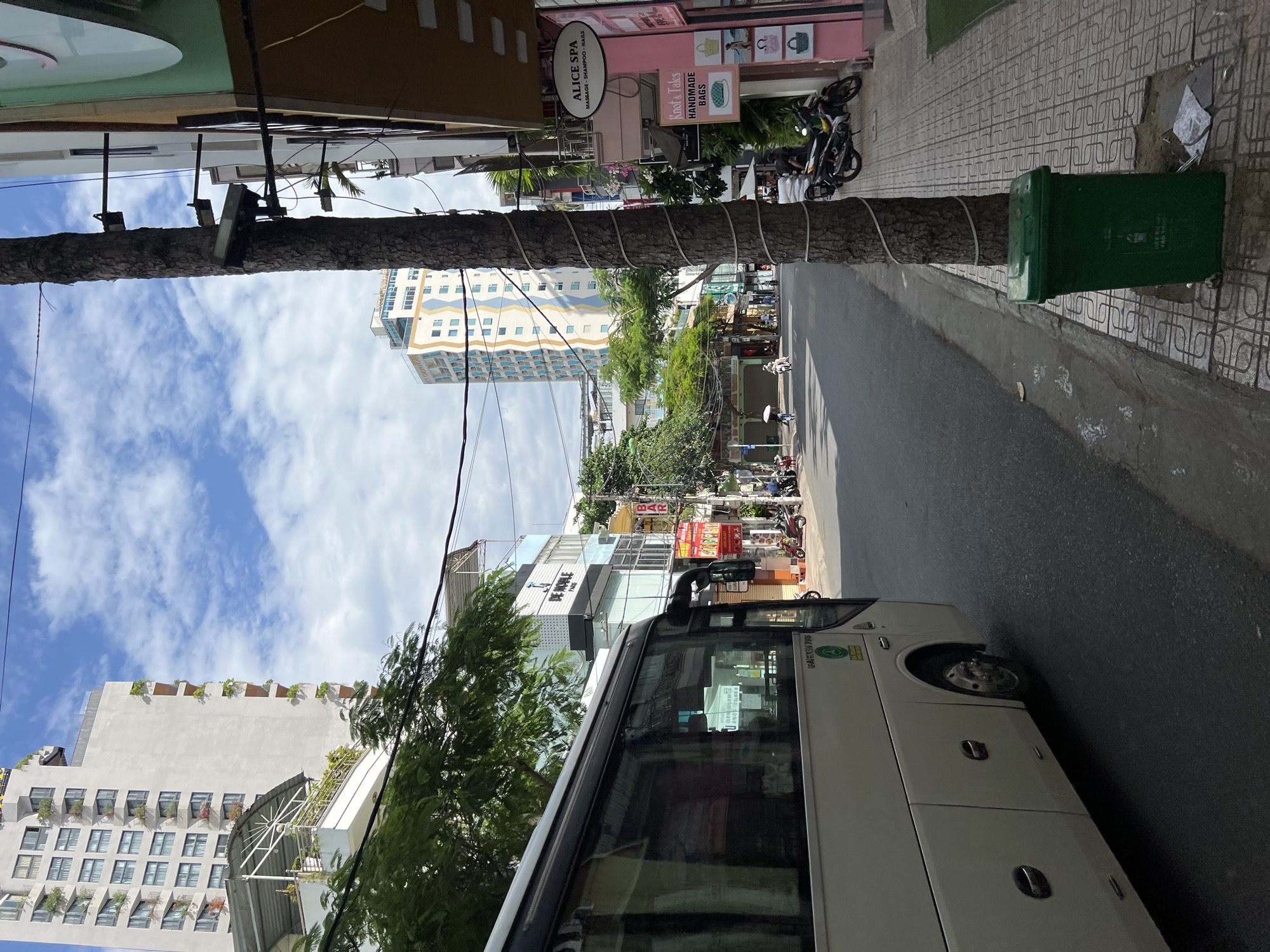 A wide shot captures a street scene in Nha Trang, Vietnam, on a partly cloudy day. In the foreground, the side of a white bus is visible, facing left. The street is paved, with a pedestrian sidewalk on the right. A tall, mature tree with rough bark stands on the sidewalk, a green trash bin is placed at its base. The sidewalk is made of patterned tiles, and several motorcycles are parked along the buildings.
On the right side of the street, buildings are lined up, featuring various businesses. Signs for "ALICE SPA MASSAGE - SHAMPOO - NAILS" and "Knot & Tales HANDMADE BAGS" are prominent. Further down the street, more buildings are visible, including one with a blue and white wavy facade. In the distance, a tall, light-colored skyscraper with many windows dominates the skyline. People are seen walking and riding motorcycles on the street and sidewalk. Lush green trees are interspersed throughout the urban landscape, providing a touch of nature against the backdrop of city architecture. The sky is a mix of blue with scattered white clouds.