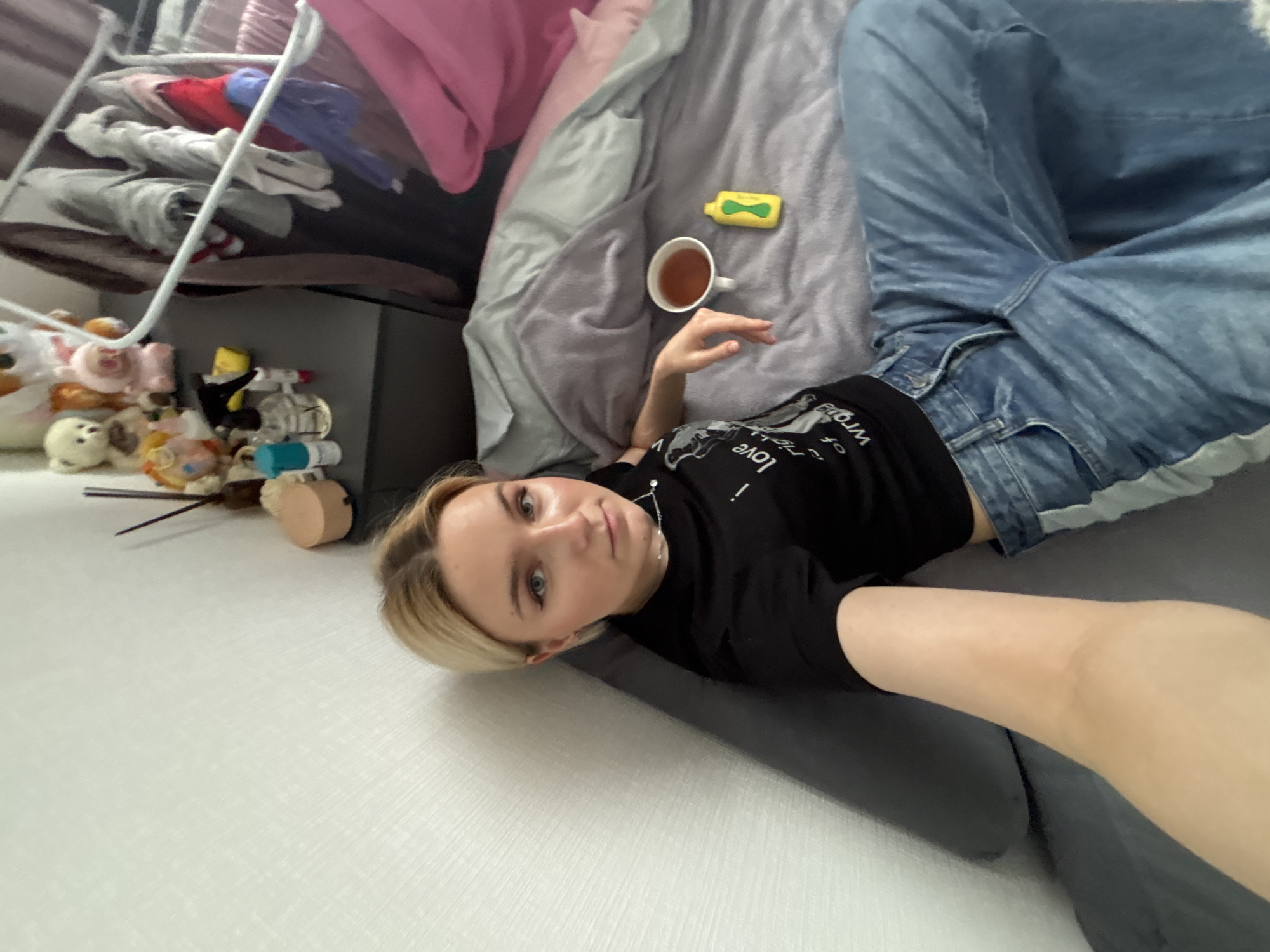 A young adult woman lies on her back on a grey blanket in what appears to be a residential room. She is wearing a black t-shirt with white writing on it and light blue jeans. Her blonde hair is pulled back. A white mug containing a dark liquid and a yellow object rest on the blanket near her. A shelf containing clothes and toys is visible in the background. The location is unknown, Russia.
