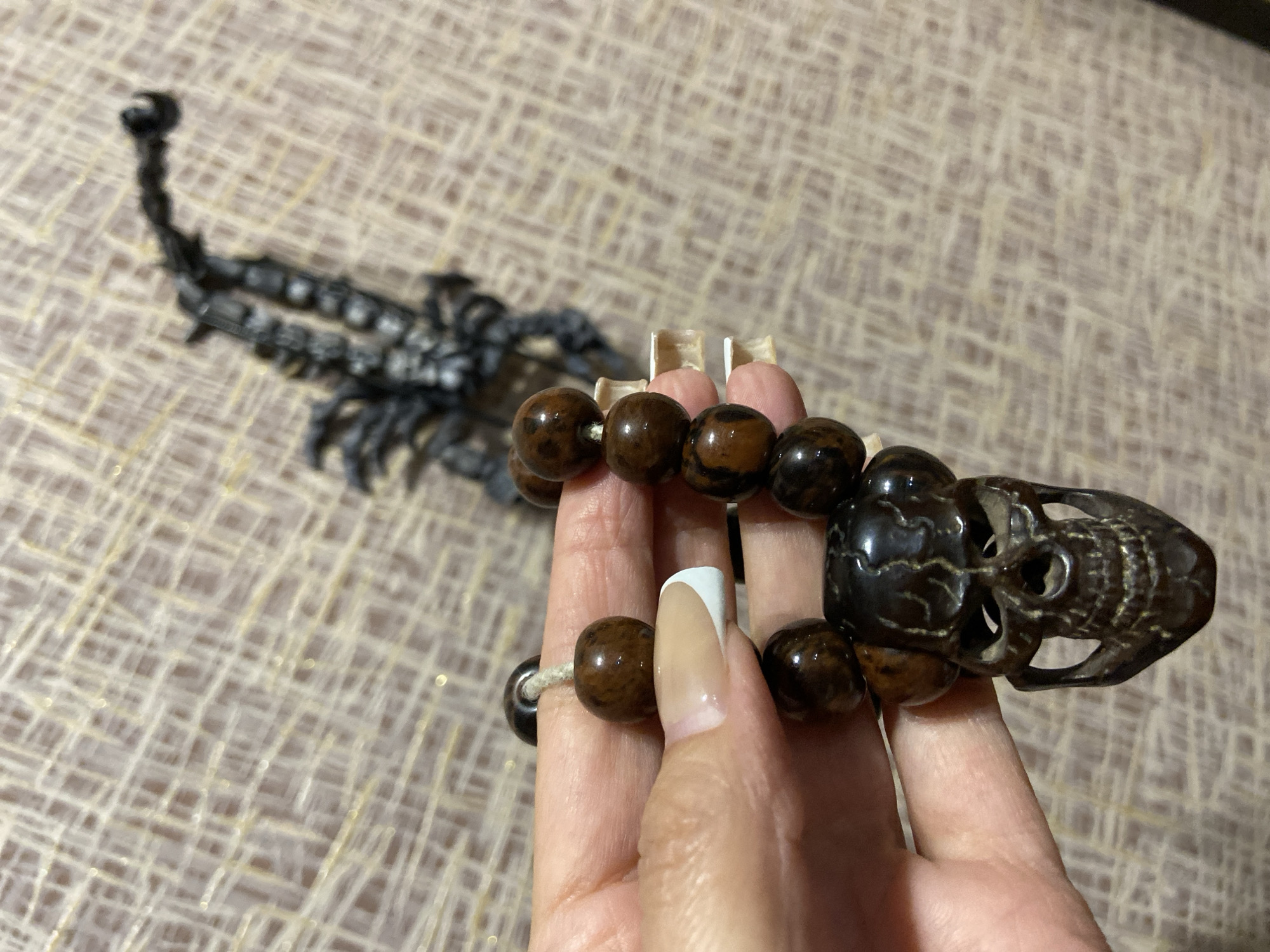 The image depicts a close-up of a right hand, manicured with a French tip style (nude base with white tips), holding a dark brown beaded item. This scene is situated in the city of Tuymazy, Russia. The spherical beads are glossy, exhibiting variegated dark and reddish-brown tones, strung on a light-colored (off-white) cord. Suspended from the beads is a prominent, dark brown, intricately carved charm shaped like a human skull, featuring defined eye sockets, nasal cavity, and individual teeth. The background is an out-of-focus, light brown or beige textured surface, possibly fabric or wallpaper, with a criss-cross pattern of lighter, reflective threads. In the upper right background, an indistinct, dark, highly textured object resembling a scorpion or a skeletal form is partially visible.