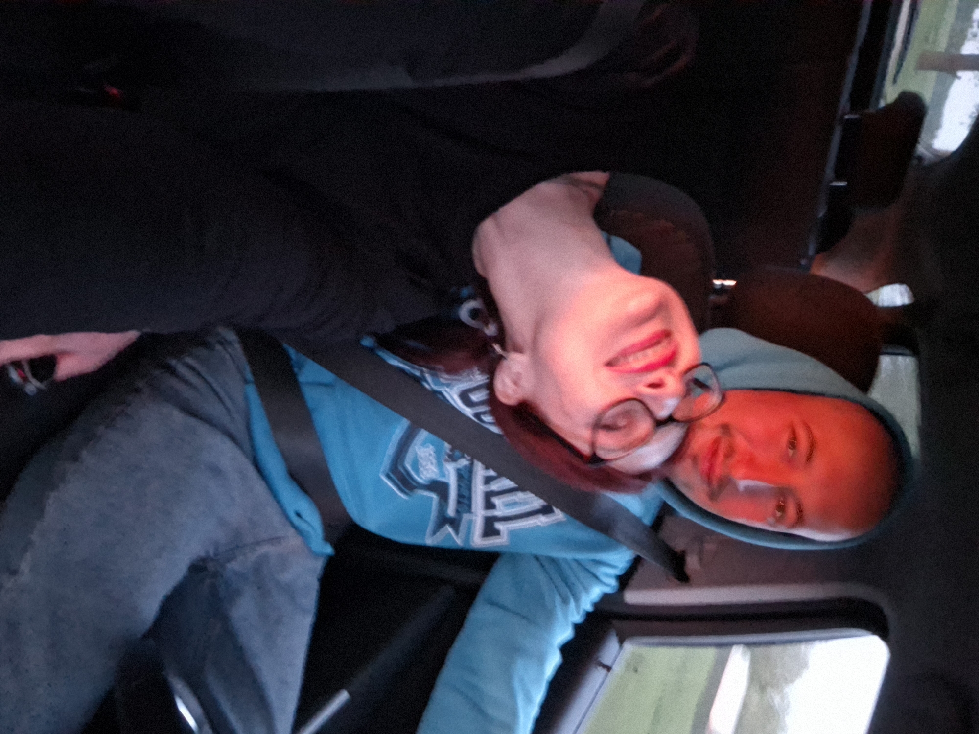 A woman wearing glasses and a blue hooded sweatshirt is sitting in a car with another person. The woman is wearing blue jeans and has a seatbelt fastened. The location is Unknown, Unknown.