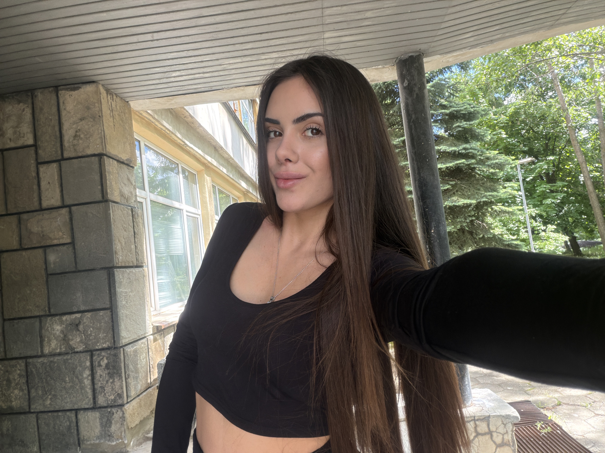 A woman is taking a selfie outdoors, standing under a covered area with a concrete ceiling. The woman has long, dark brown hair and is wearing a black long-sleeved crop top with a necklace. Part of her midriff is visible. The background includes a stone brick wall, a window, a dark gray column, and green trees. The location is likely in the city of Sofia, Bulgaria.