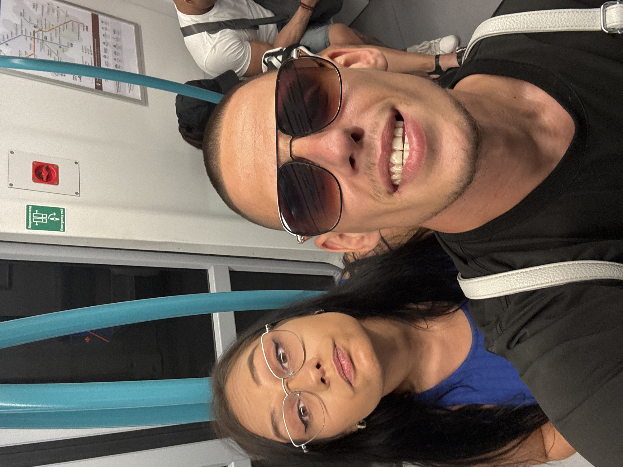A man and a woman are taking a selfie inside a subway car in Sofia, Bulgaria. The man in the foreground is wearing sunglasses and a black t-shirt, and he is smiling. The woman is to his left, wearing glasses and a blue top. She is looking at the camera with a neutral expression. The subway car has blue handrails and a map on the wall. An "Emergency Exit" sign is visible on the wall.