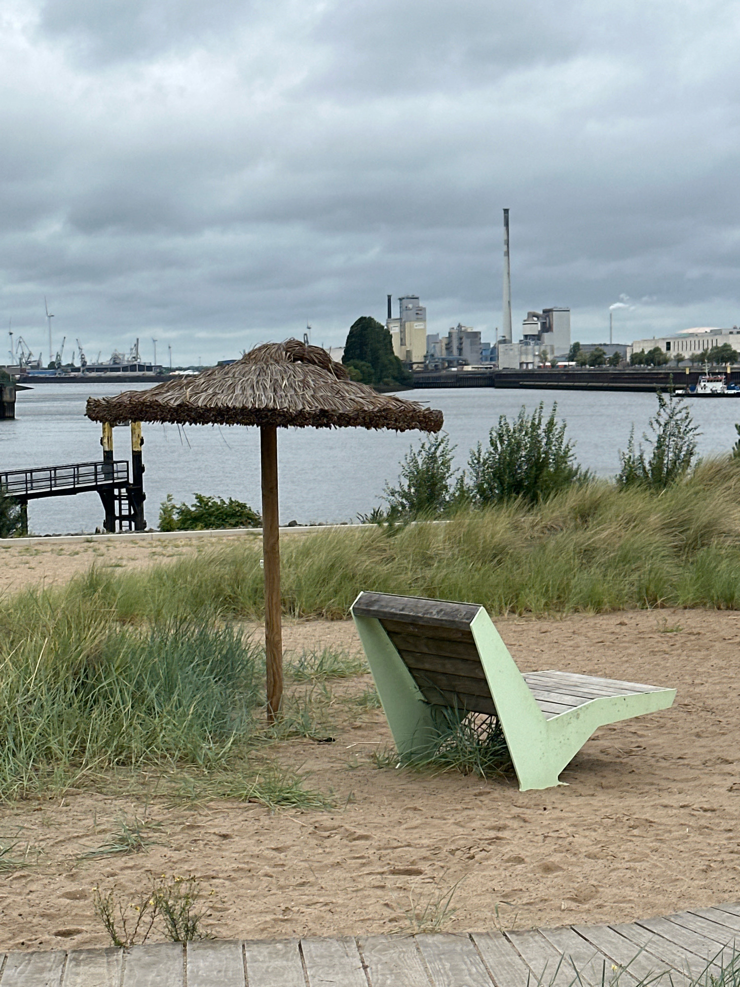The image depicts a sandy area with sparse vegetation in the foreground, featuring a wooden slatted walkway at the bottom. A light green bench with a wooden seat is positioned on the sand, angled to the right. Behind the bench and slightly to the left is a thatched umbrella with a wooden pole. In the midground, there is a body of water, likely a harbor or river, with a wooden pier and walkway extending into it on the left. Across the water, a cityscape is visible, characterized by industrial buildings with smokestacks, cranes, and wind turbines. This appears to be a view of the Bremen port area, specifically the industrial zones. The sky is overcast and gray. There are no people visible in the scene.