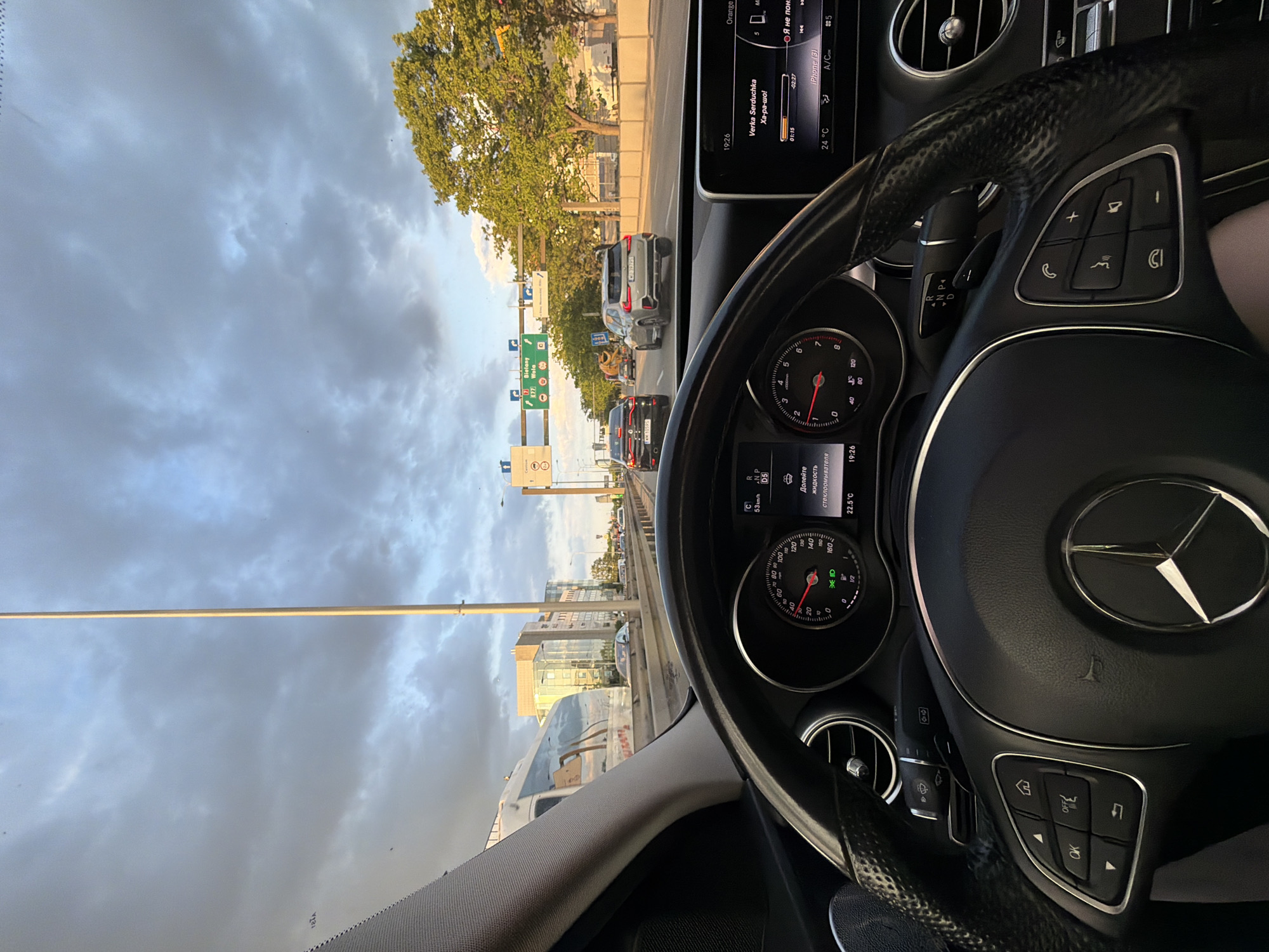 The image depicts the view from inside a Mercedes-Benz car while driving on a highway. The car is in drive mode, indicated by "D5" on the dashboard. The speed is 53 km/h. The dashboard also indicates a temperature of 22.5°C and a low windshield washer fluid level. The car stereo is playing "Verka Serduchka - Ха-ра-шо!". The highway signs visible through the windshield indicate directions to "Bielany", potentially placing the location near or in Bielany. The sky is overcast with cloudy conditions. Other cars are visible on the highway. Buildings are visible in the distance, along with trees lining the road.