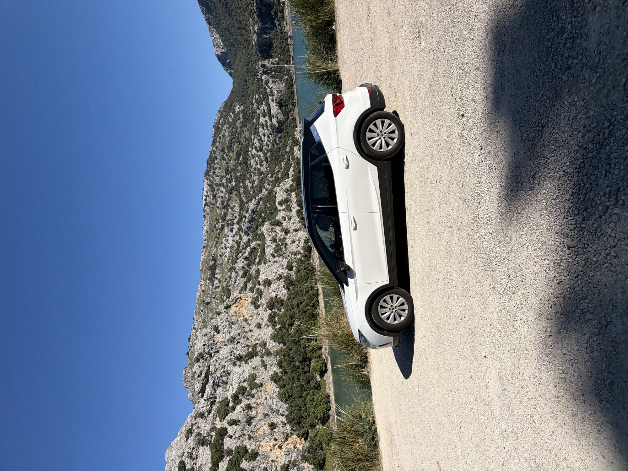 A white Opel Crossland sits on a gravel roadside in Escorca, Spain. The car is oriented diagonally in the frame. Behind the vehicle is a rugged, rocky mountain terrain interspersed with dense vegetation. A body of water, possibly a lake or reservoir, is visible at the base of the mountains. The sky above is clear and blue. The lighting suggests a sunny day, with shadows cast by the car on the gravel surface. There are no people visible in the image.