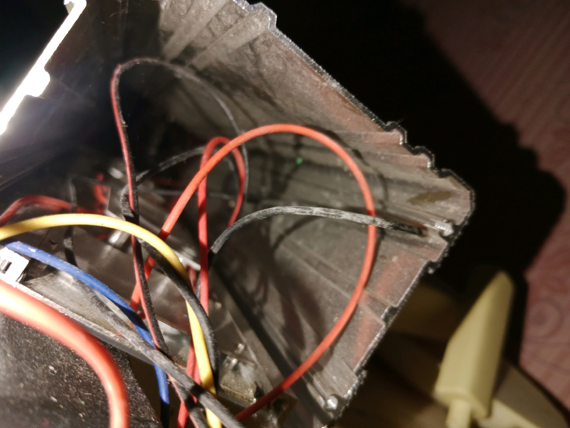 The image shows a close-up view of the interior of an electronic device, possibly a flashlight, with exposed wiring. Several wires of different colors, including red, yellow, blue, and black, are visible, some of which appear to be frayed or damaged. The casing of the device is made of a dark, metallic material with a textured surface. A bright light source is illuminating the scene from the left side of the image, casting shadows. A beige-colored plastic component is visible in the lower right quadrant of the image, partially out of focus. The overall environment appears to be indoors, with a textured surface, possibly a table, visible in the background. There are no people or animals present in the image. The specific location in Ringinrejo, Indonesia, is not discernible from the visual information provided.
