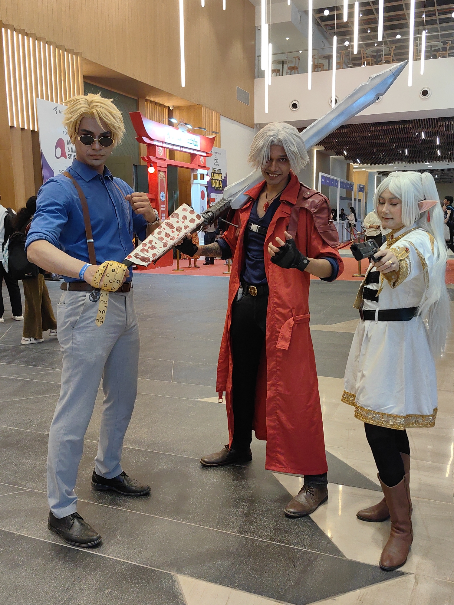 The image depicts three individuals dressed in elaborate cosplay costumes at what appears to be an indoor event in Mumbai, India. In the foreground, from left to right, a person with blonde, spiky hair wearing sunglasses, a blue collared shirt with brown suspenders, light grey pants, and black dress shoes is holding a prop resembling a large, stylized katana. To their right, a person with white, shoulder-length hair wearing a red trench coat over a dark shirt and black pants, holding a large silver sword over their shoulder, is looking towards the camera with a smile. To the right of the sword-wielder, a person with long, white hair, wearing a white and gold outfit with pointed ears and brown boots, is holding a black prop gun and looking to their left. The background suggests a convention or exhibition hall with an architectural style featuring vertical lighting elements and wooden accents. A red torii gate structure with Japanese text is visible behind the leftmost individual. The floor is tiled with a geometric pattern of dark grey and lighter grey tiles. Signs in the background, including one with "ANIME INDIA," indicate the event's theme. The overall scene suggests a lively cosplay event.