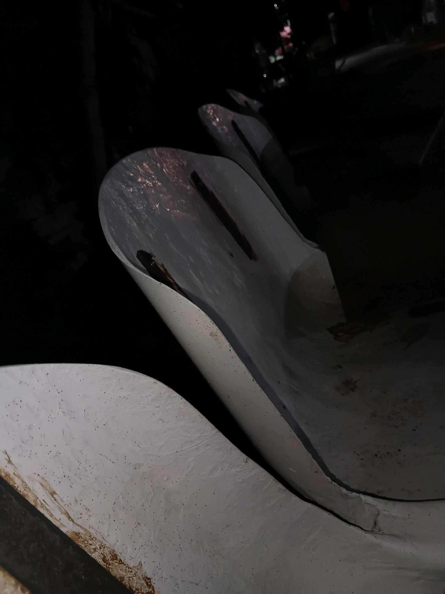 The image shows a close-up, low-angle view of two white, curved structures, possibly seating or structural elements, in what appears to be an outdoor urban setting at night. The surfaces are textured and show some signs of wear and tear, with dark spots and a slight sheen in places. In the background, blurred lights suggest the presence of buildings and possibly streetlights, indicating an urban environment. The context of Cikal, Indonesia, is not discernible from the visual information presented in the image itself.
