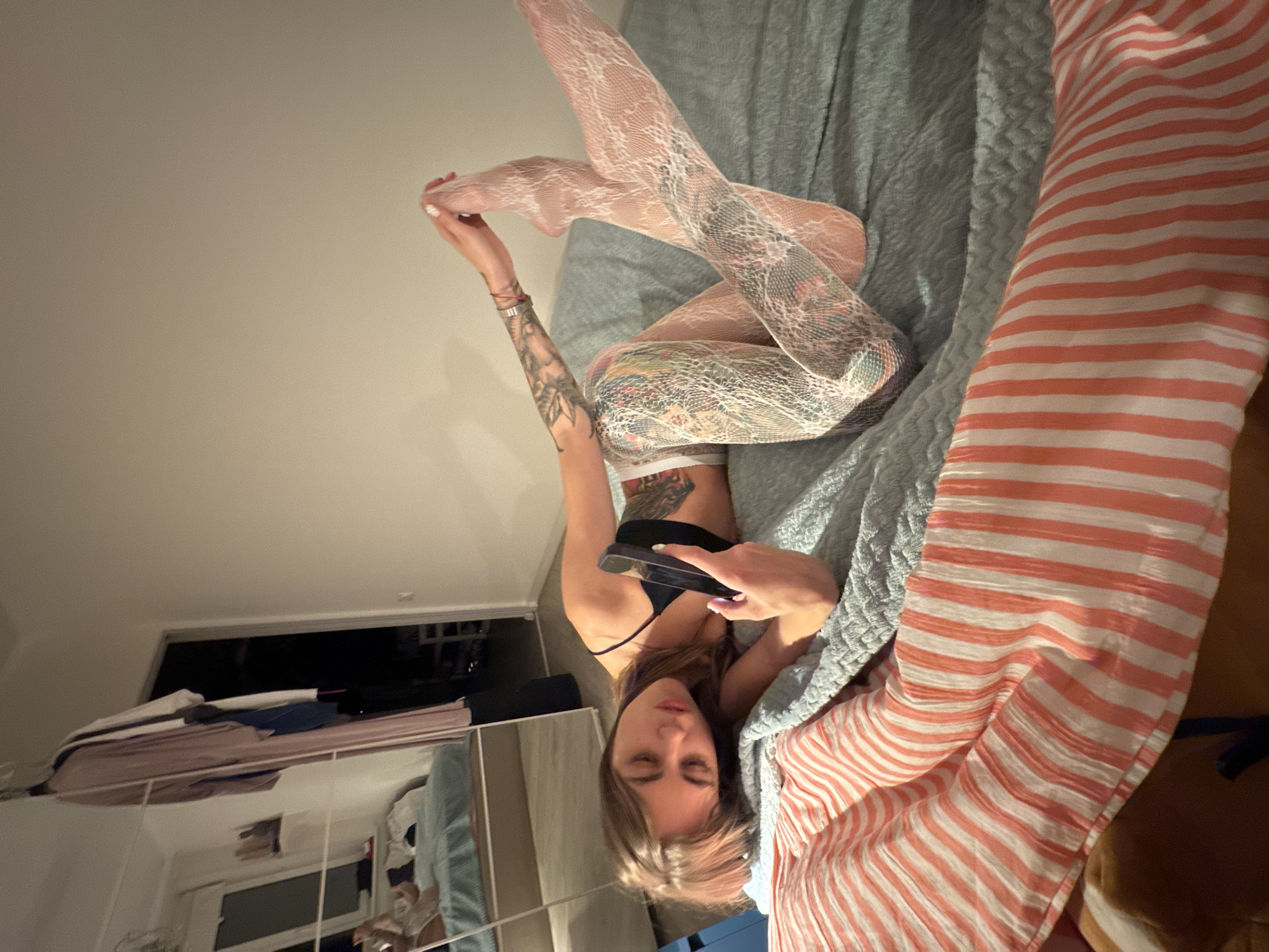A young woman with tattoos on her arm and leg is lying on a bed, holding a smartphone. She is wearing a black bra and white lace stockings. The room appears to be a bedroom, with a closet in the background containing clothes on hangers. The bed is covered with a light blue textured blanket and a striped comforter. The lighting suggests it is evening or night. There are no clear indications of the location being specifically in Moscow, Russia.