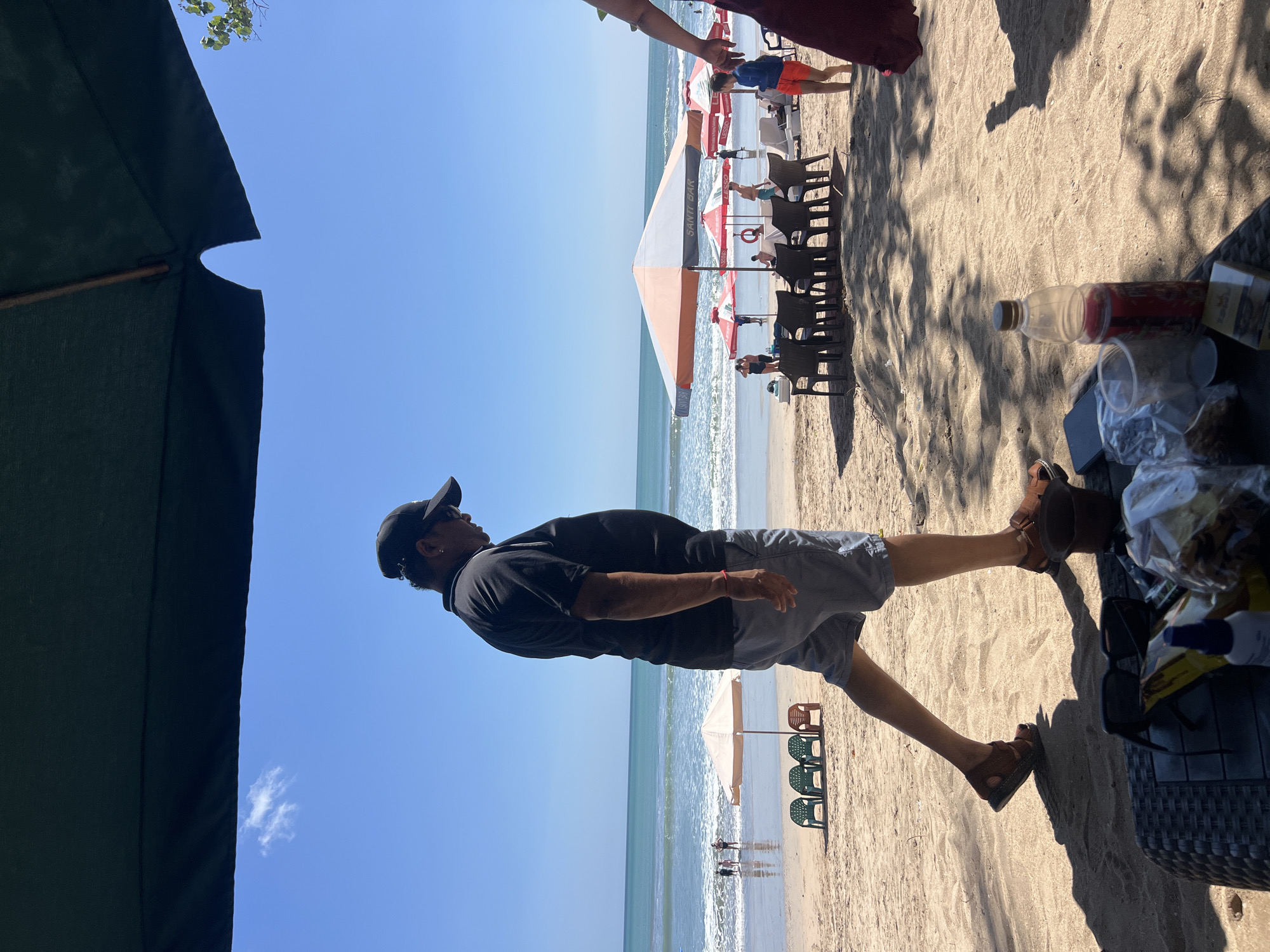 This image depicts a beach scene in Kuta, Indonesia, on a bright, sunny day. A man in a black t-shirt, grey shorts, and a black baseball cap walks across the sand in the foreground, casting a shadow. He is wearing brown sandals. In the middle ground, several beach chairs and umbrellas are set up on the sand. The ocean with gentle waves is visible in the background, with a few people in the water. Another person, only partially visible from the side, is walking away from the camera on the right side of the frame. On a table in the lower right corner, there are sunglasses, a bottle of liquid, and some packaged items. The overall scene conveys a relaxed atmosphere typical of a beach resort.