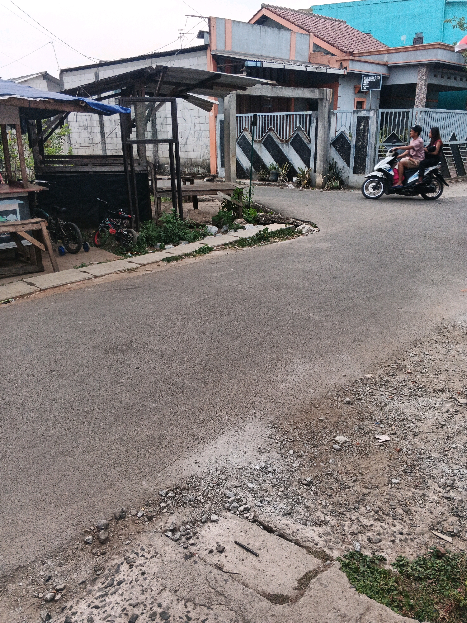 A scene unfolds in Kadu, Indonesia, with a paved road curving through a residential area. On the left, a makeshift stall with a blue tarp roof stands next to wooden structures. Two bicycles, one with training wheels, rest near the stall. In the background, a concrete fence with a decorative pattern divides properties, and houses with various roof styles and colored facades, including a bright blue one, are visible. On the road, two individuals ride a white motorcycle. The driver, wearing a pink shirt and a black cap, is accompanied by a passenger in a dark top. The road's edge is bordered by gravel and sparse vegetation. The sky above is overcast.