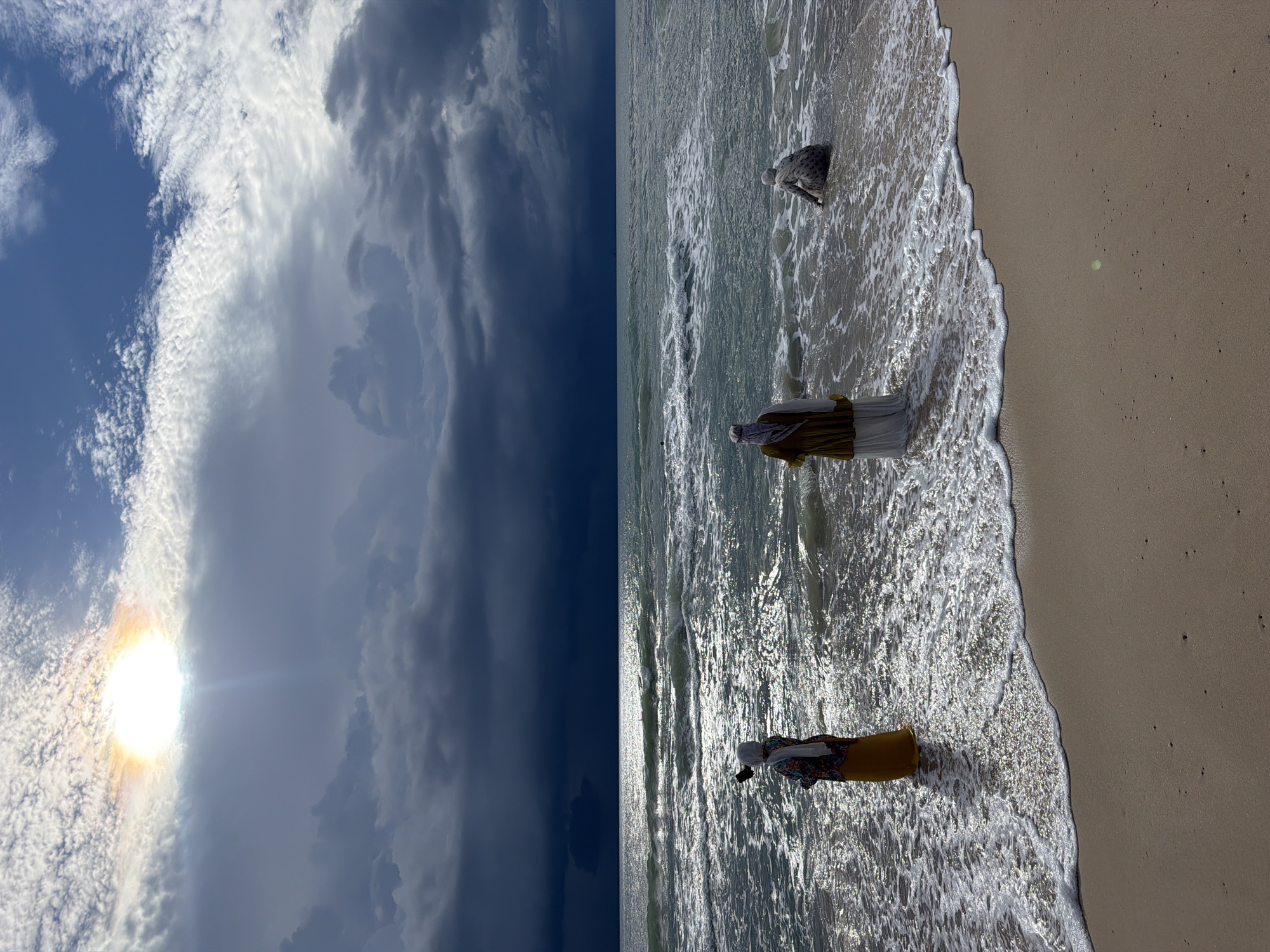 The image depicts a beach scene with three individuals near the shoreline. The sky is partially cloudy with the sun visible through a break in the clouds, casting a bright glare. The sea is choppy with waves approaching the shore. The sand in the foreground is light brown.
On the left, a person wearing a hijab and a brightly colored patterned top over a yellow skirt stands in shallow water, holding a mobile phone as if taking a selfie. In the center, another person, also wearing a hijab and a long, light-colored garment, stands in the shallow water facing the sea. To the right, a third person wearing a patterned outfit and a hijab is crouched down at the edge of the water, partially submerged. The individuals appear to be women.
The overall atmosphere suggests a dramatic interplay of light and dark, with the sunlit sky contrasting with dark, ominous clouds over the ocean. The location is not identifiable as being in Choeng Thale, Thailand, based on visual information in the image.