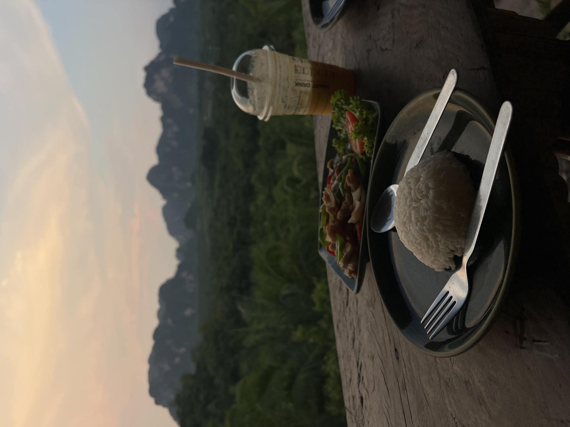 The photograph shows a meal served on a wooden table against a backdrop of mountains and greenery, possibly in the Khao Thong area of Thailand. The meal consists of a plate of white rice with a fork and spoon, a plate of stir-fried vegetables and meat, and a plastic cup containing a beverage. The sky is visible in the background, indicating it could be either dawn or dusk.