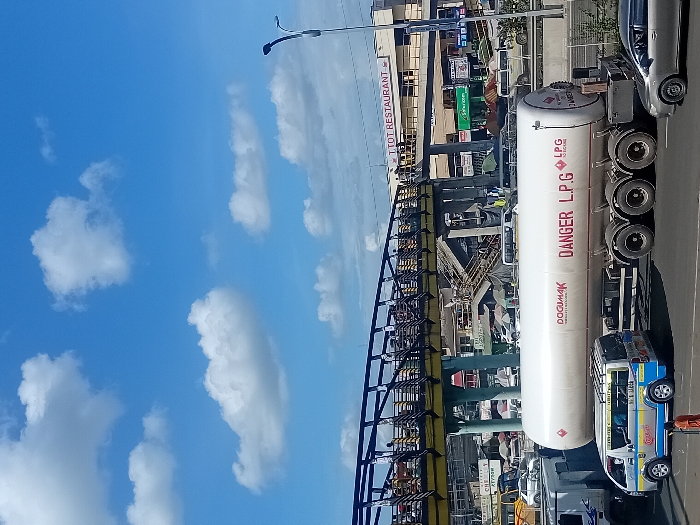 The image shows a busy street scene in Mlolongo, Kenya, located at coordinates -1.394 latitude and 36.94 longitude. A large white tanker truck, labelled "Dоgиmаk" and "DANGER L.P.G" is parked in the middle of the road. It appears to be transporting liquefied petroleum gas (LPG). Behind the truck, a pedestrian bridge crosses the street with a small number of pedestrians walking on it. In the background, a large building with a sign reading "T.O.T. RESTAURANT" is visible. The scene is framed by a clear blue sky with fluffy white clouds. Several vehicles, including a silver car, a white van, and a truck, are visible in the background. There are also several shops and stalls on the sides of the street. The image suggests a bustling commercial area with a mixture of traffic and pedestrians.